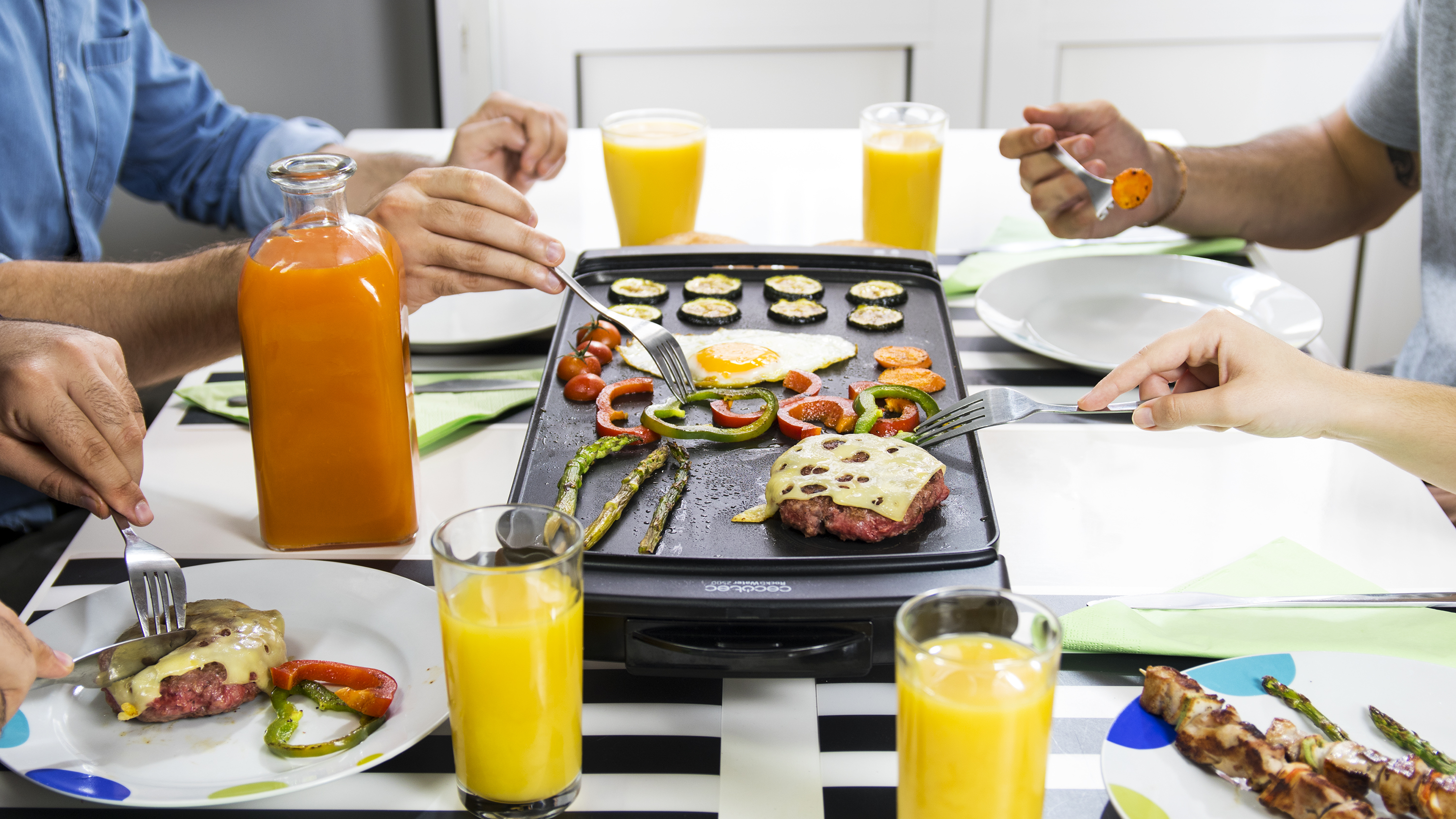 Personas comiendo en una mesa con una plancha que cocina comida. Comida a la parrilla y bebidas visibles.