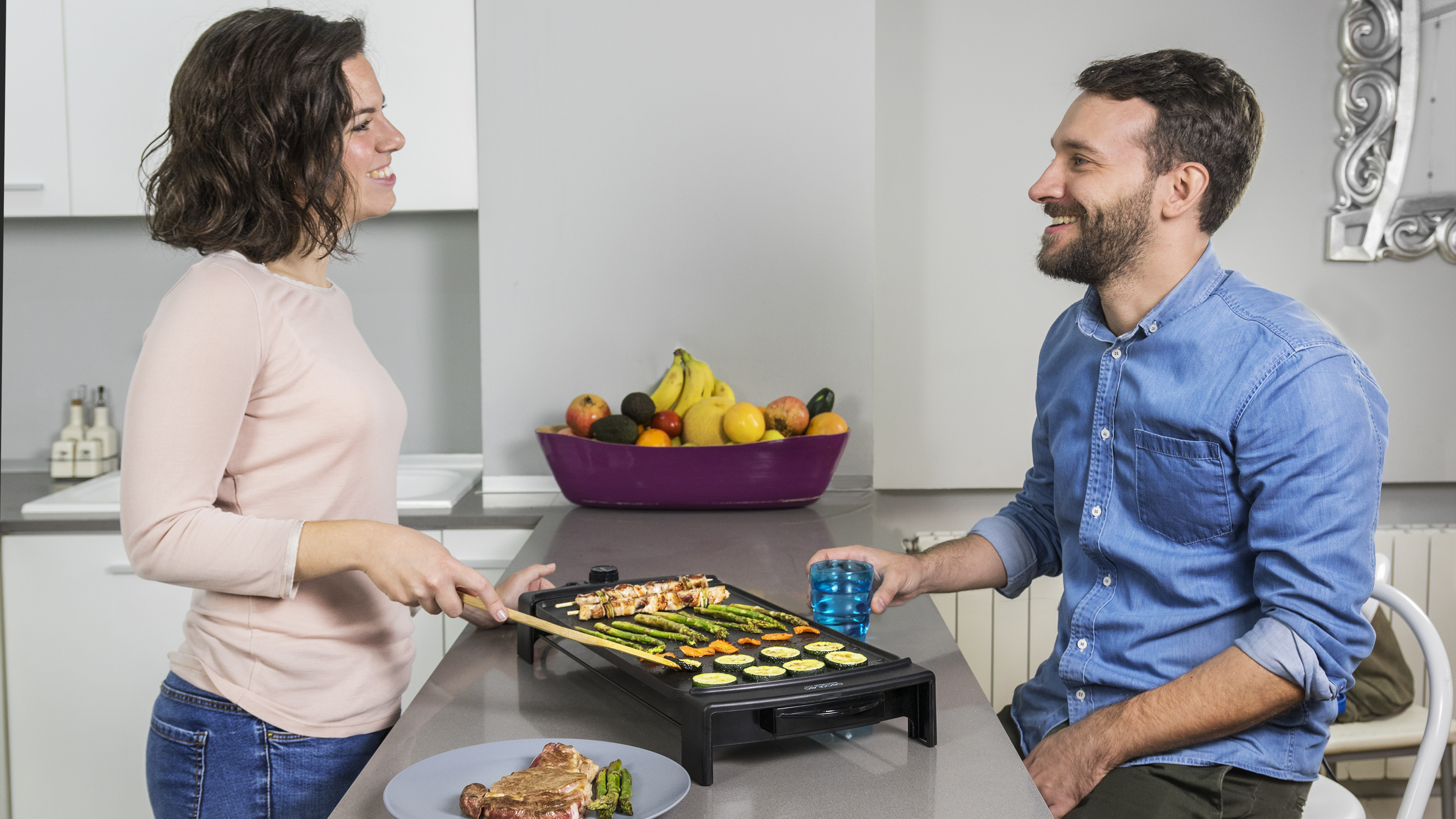 Un hombre y una mujer sonríen mientras asan comida en una parrilla de encimera, en una cocina.