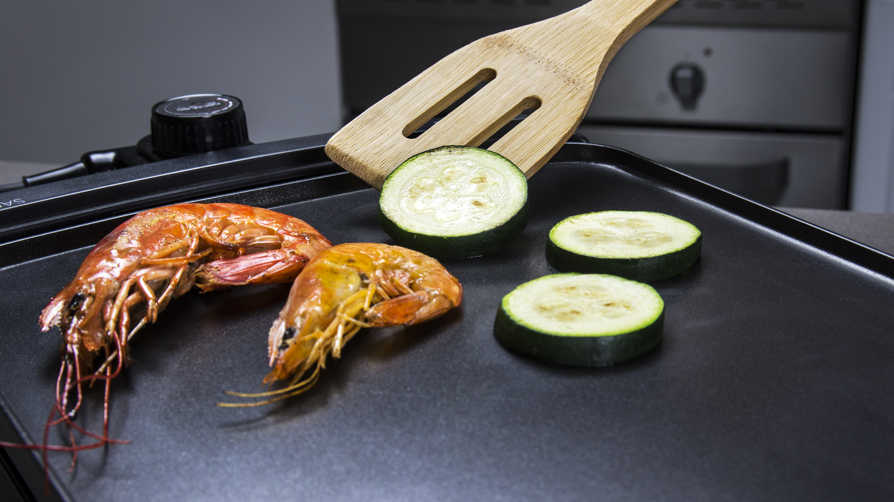 Camarones y rodajas de calabacín en una plancha negra con una espátula de madera.