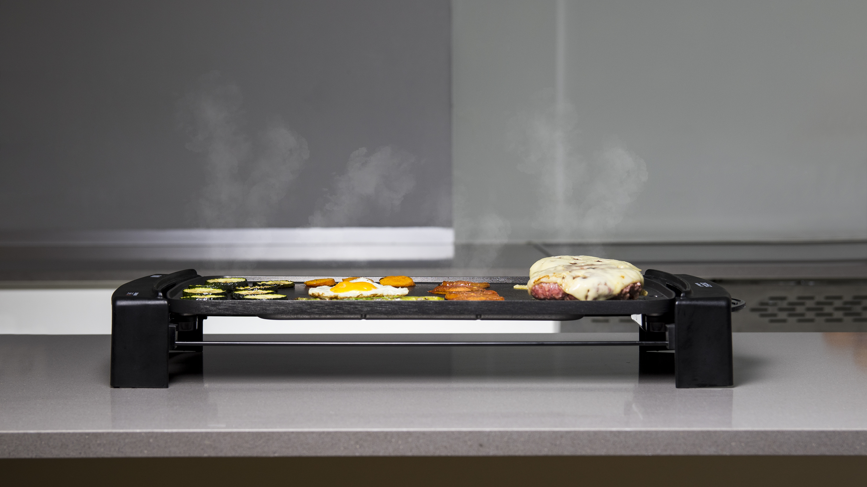 Una parrilla eléctrica con comida cocinándose, incluyendo vegetales y carne, con vapor subiendo contra un fondo gris.
