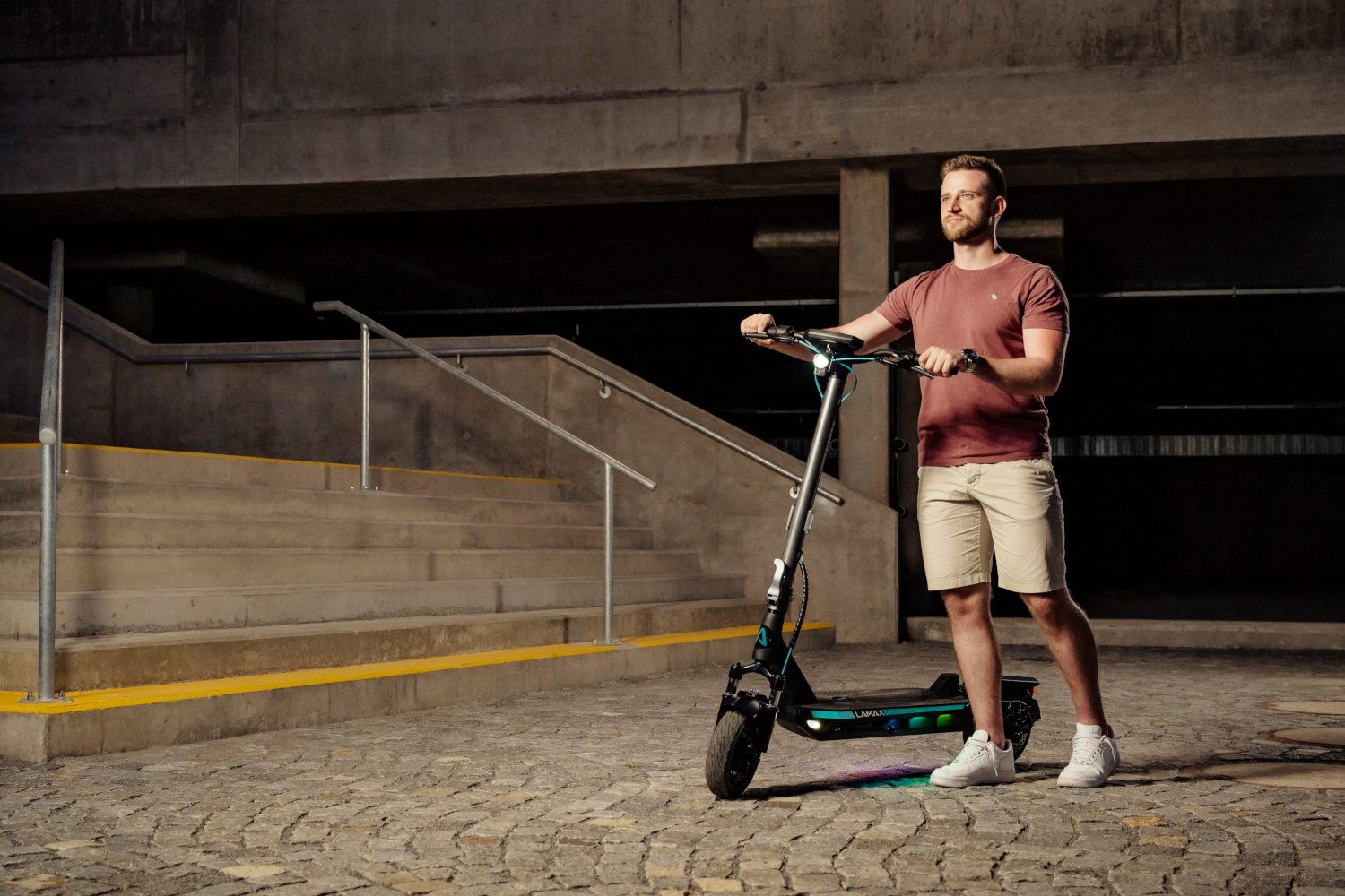Férfi elektromos rollerrel betonon áll. Mögötte lépcsők és egy sötét parkoló.