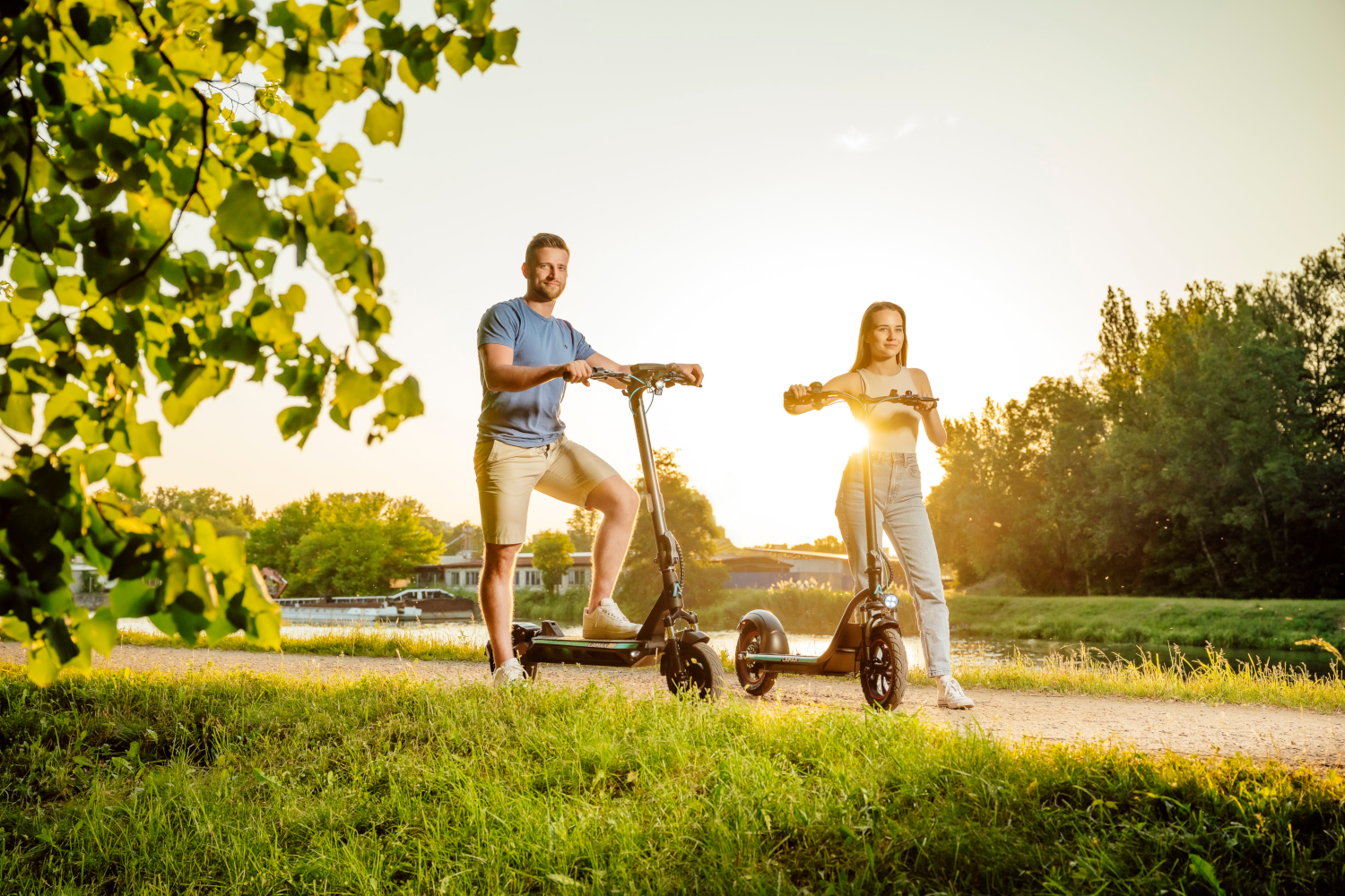 Két ember robogóval, egy ösvényen. Egy férfi, egy nő, mindketten mosolyognak. Zöld és fák a háttérben.