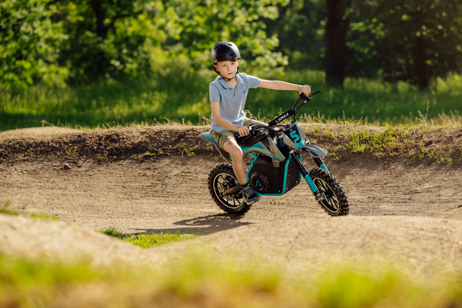 Egy fiú mini dirt bike-on versenypályán. Sisakot visel, előre néz. Napos kültéri környezet.