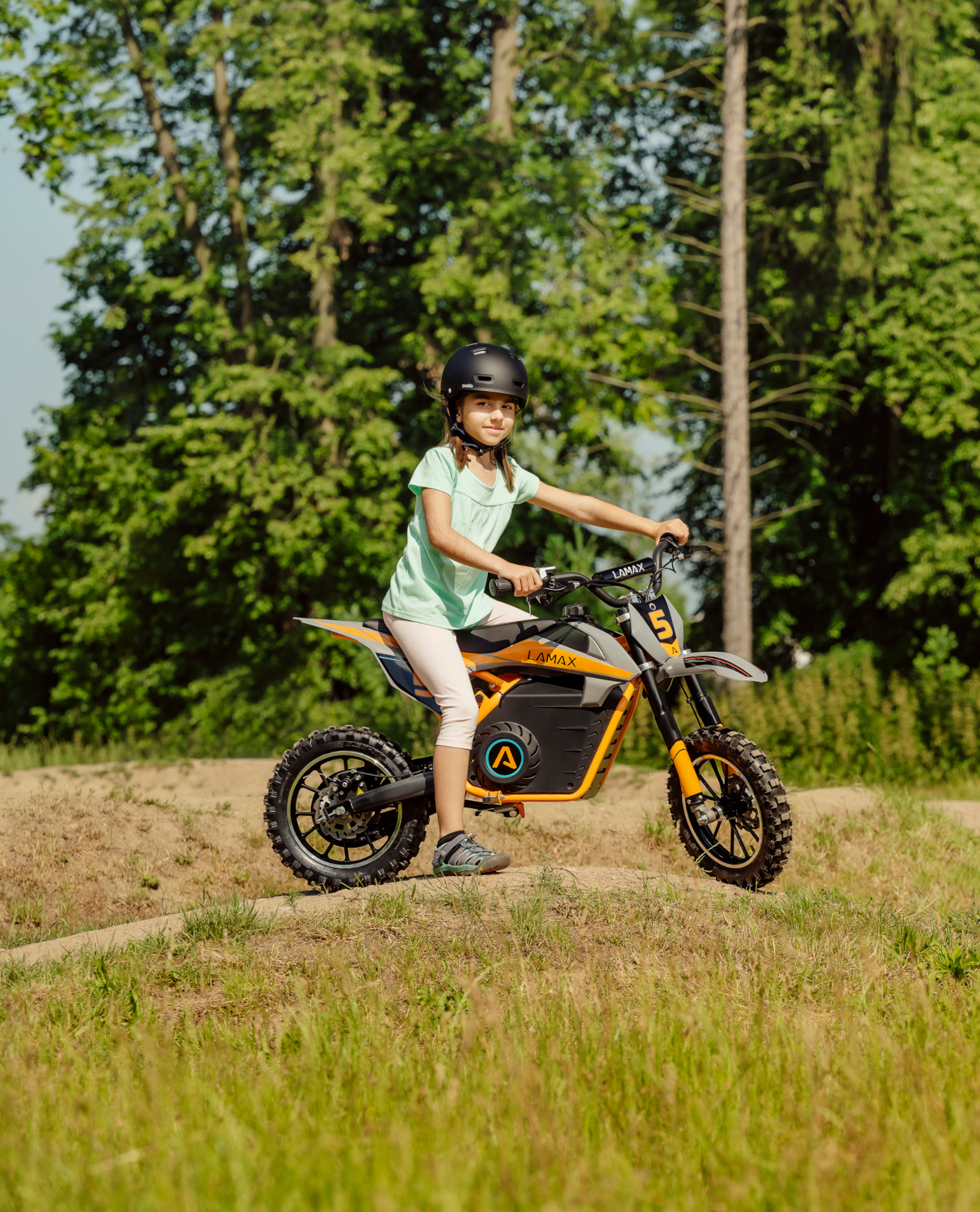 Egy lány sisakban narancssárga-fekete elektromos motorkerékpárral a füves pályán.
