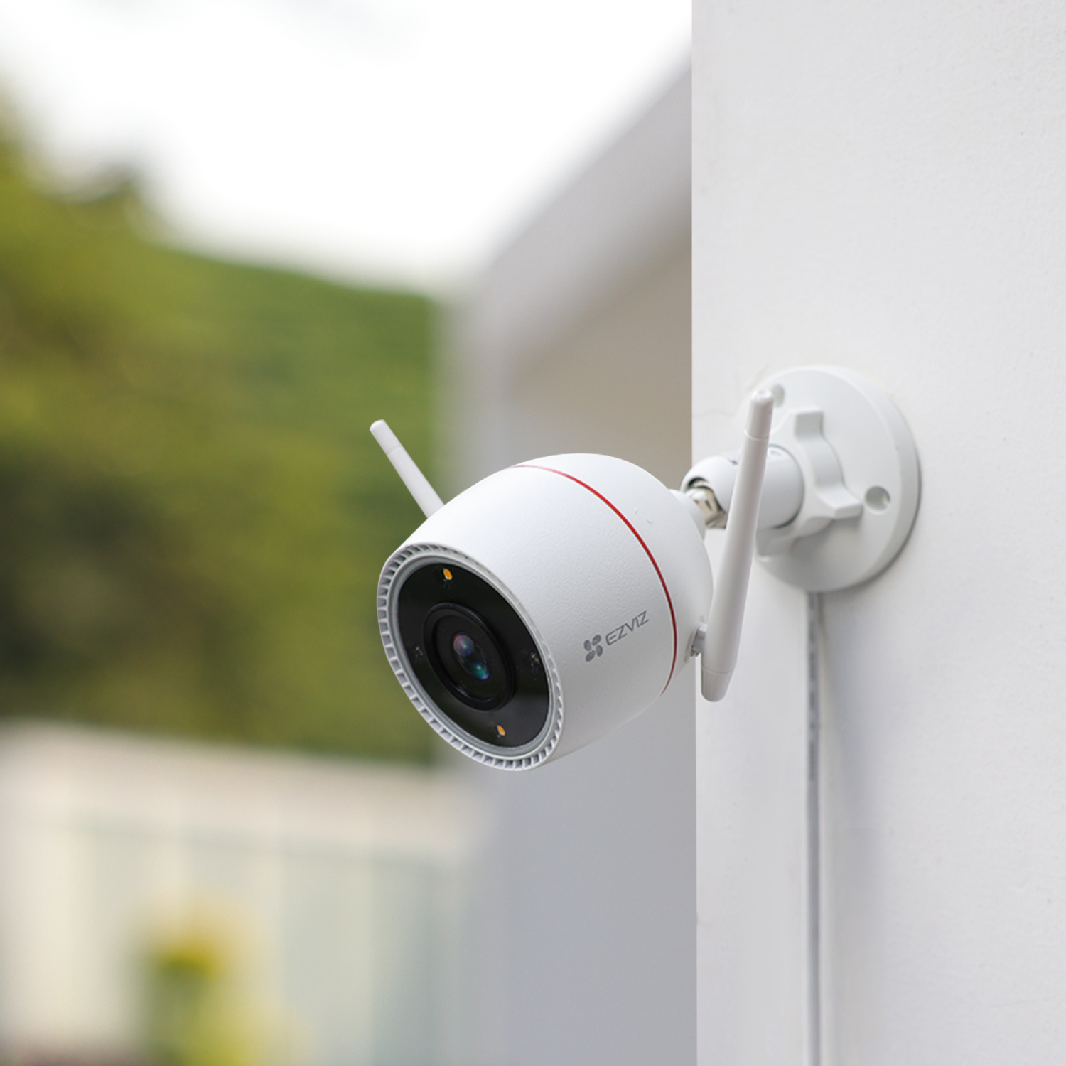 White security camera with two antennas, mounted on a white wall, outdoor scene.