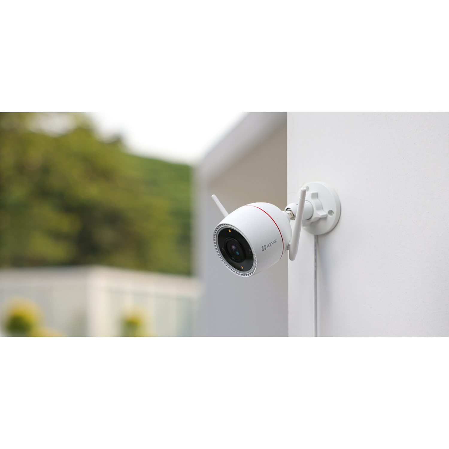 A white security camera attached to a white wall with two antennas, with the background blurred.