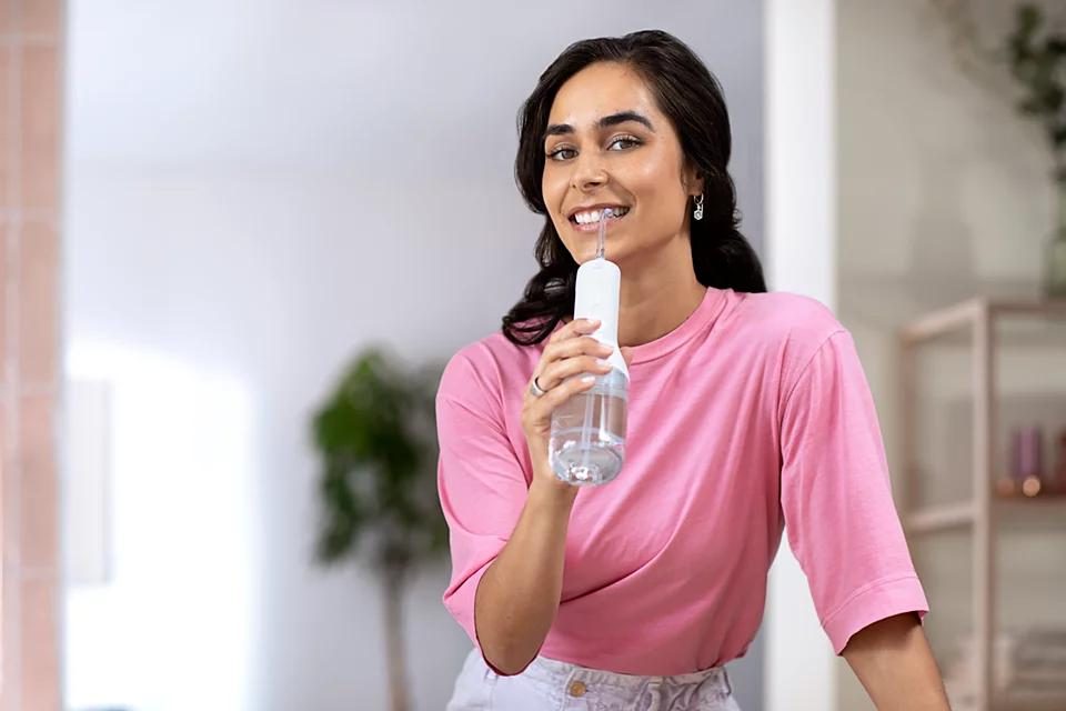 Uśmiechnięta kobieta trzymająca irygator do zębów. Ma na sobie różową koszulę.