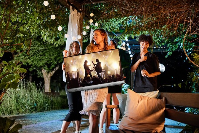 Trzy osoby trzymają telewizor na nocnej imprezie plenerowej, z lampkami choinkowymi.