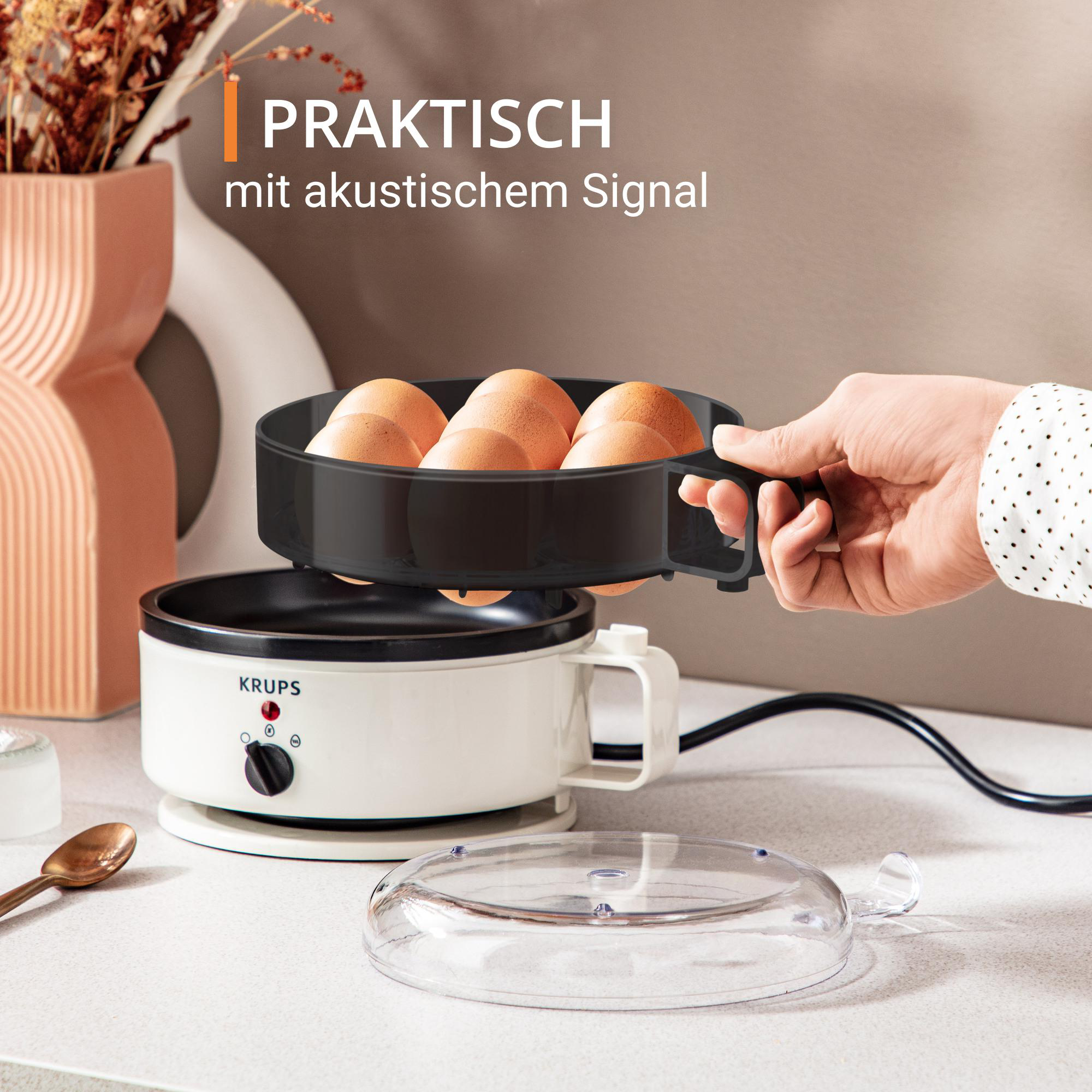 Eine Hand, die ein Eiertablett über einem Eierkocher hält, mit Eiern, Text und einem Deckel.