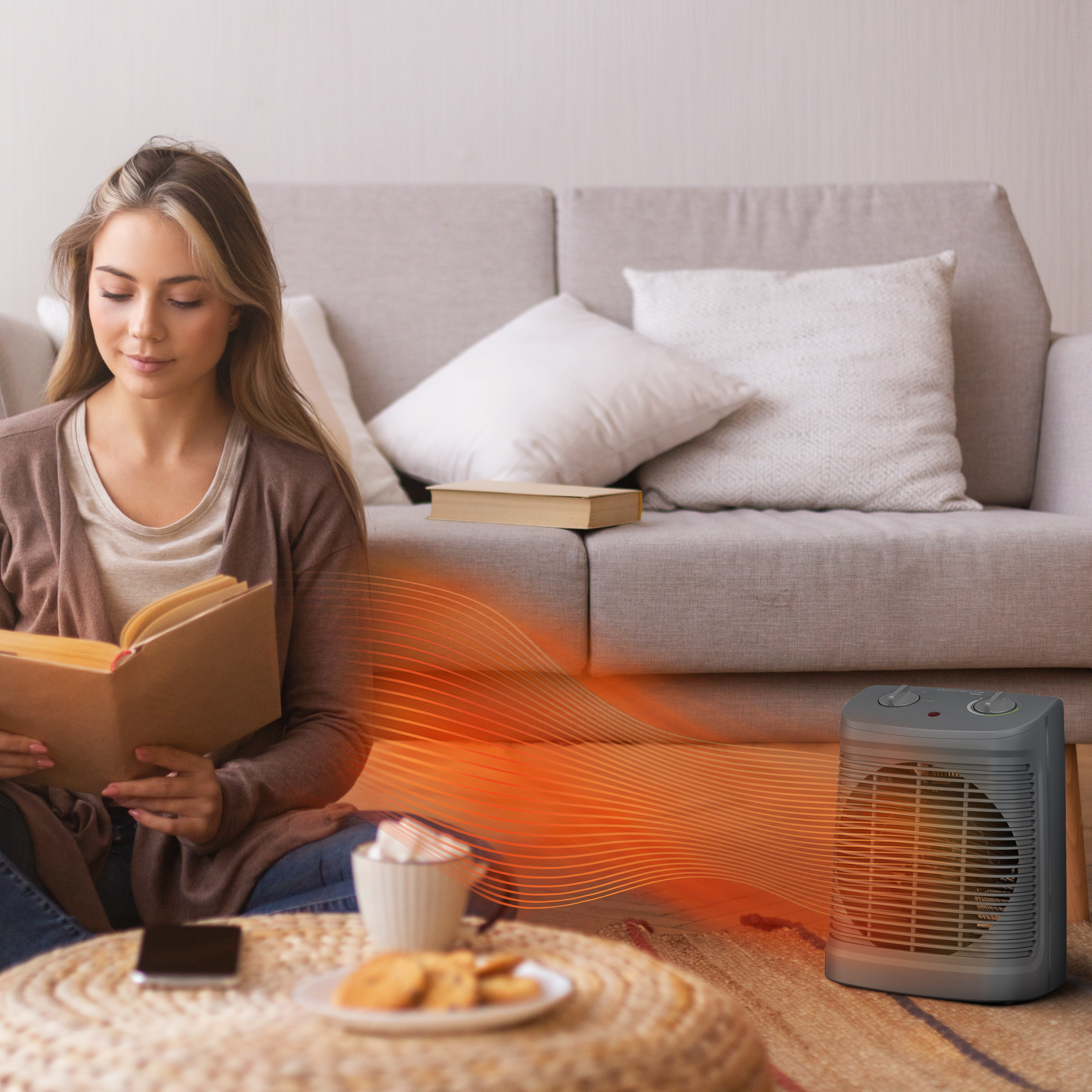 Mujer leyendo un libro junto a un calefactor. Café y galletas en una mesa. Un sofá en el fondo.