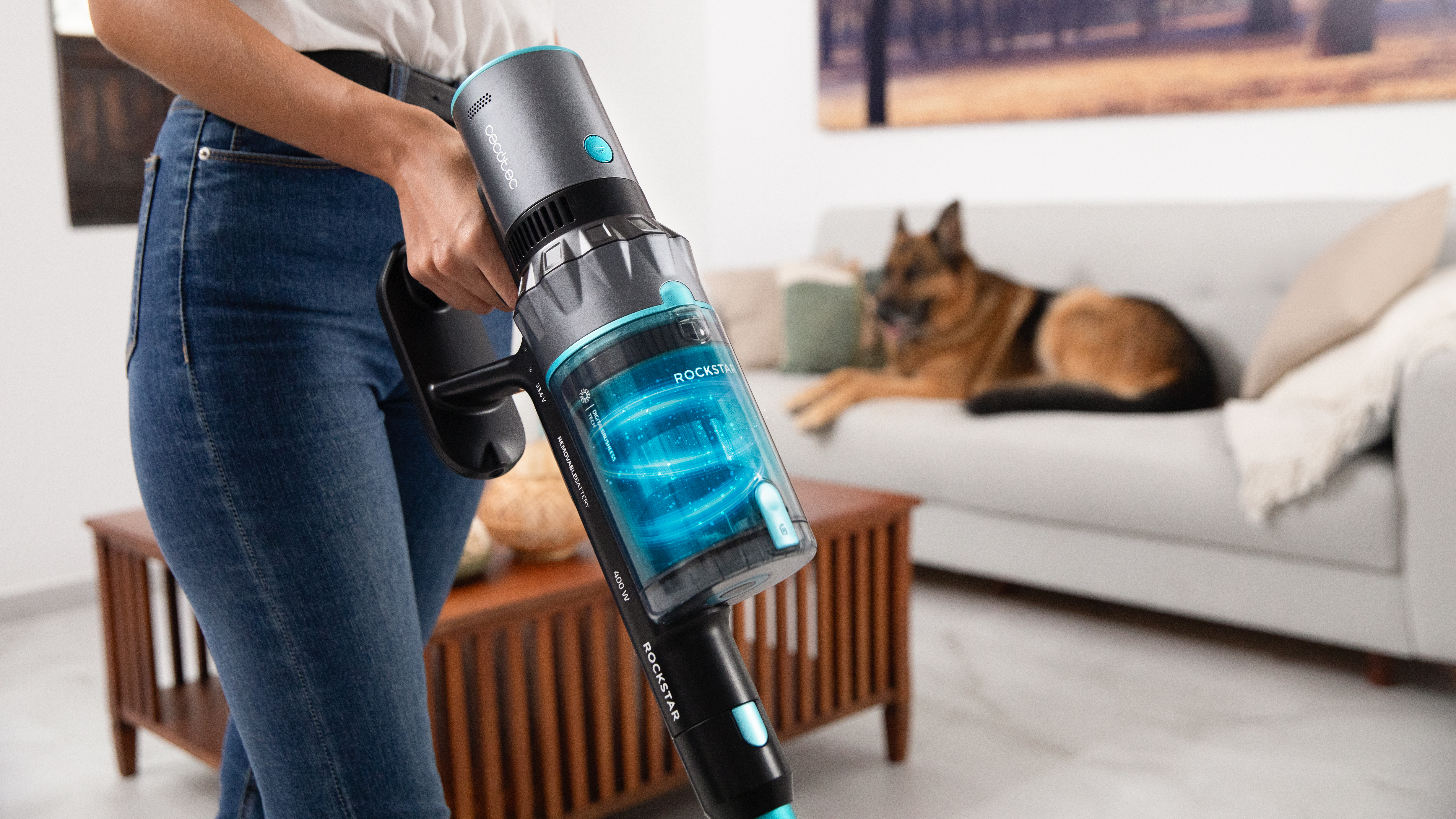 Mujer usando una aspiradora Cecotec Rockstar negra y azul. Un perro está en un sofá al fondo.