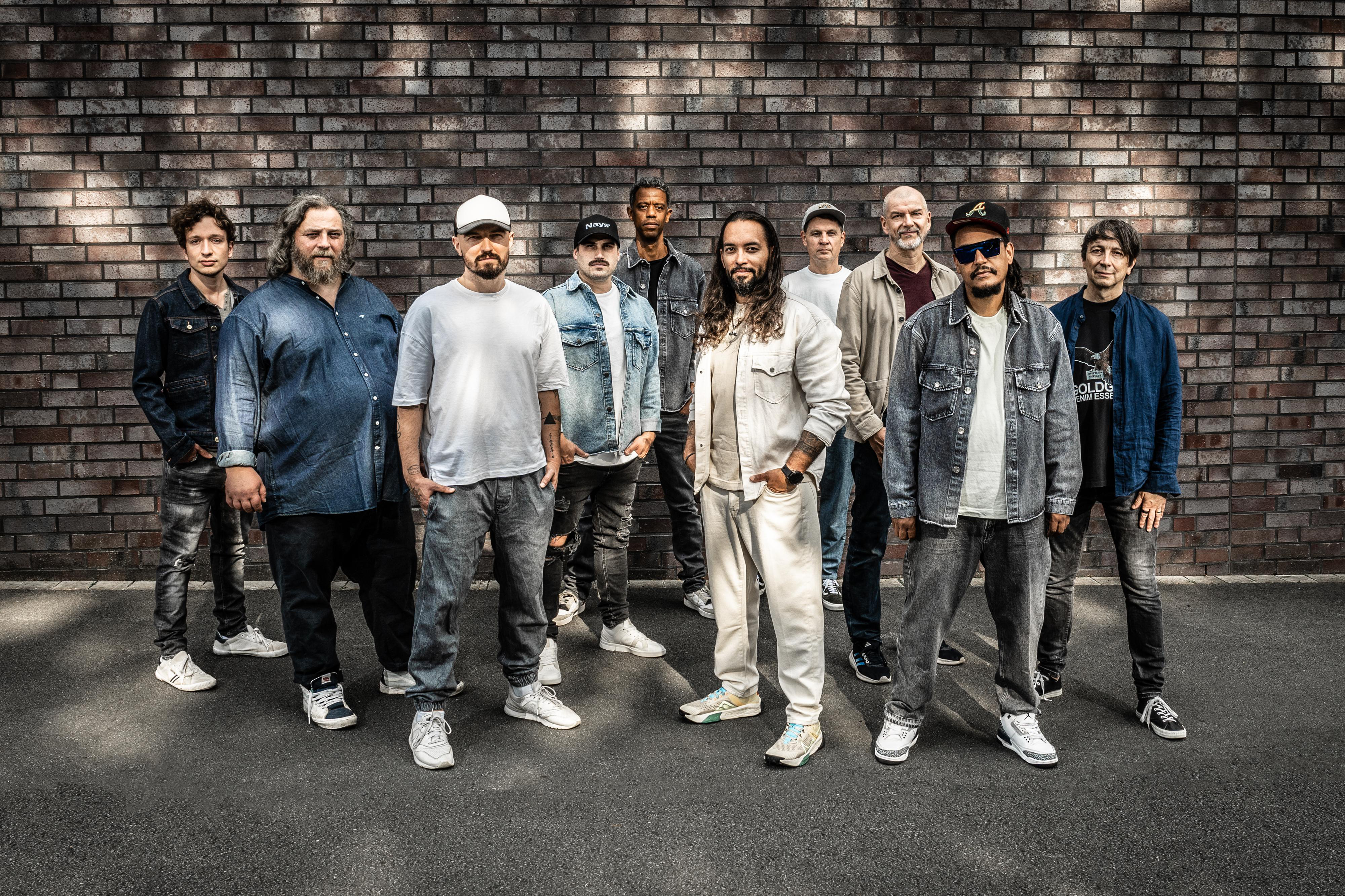 Männergruppe steht vor einer Backsteinmauer. Sie tragen alle unterschiedliche Kleidung.