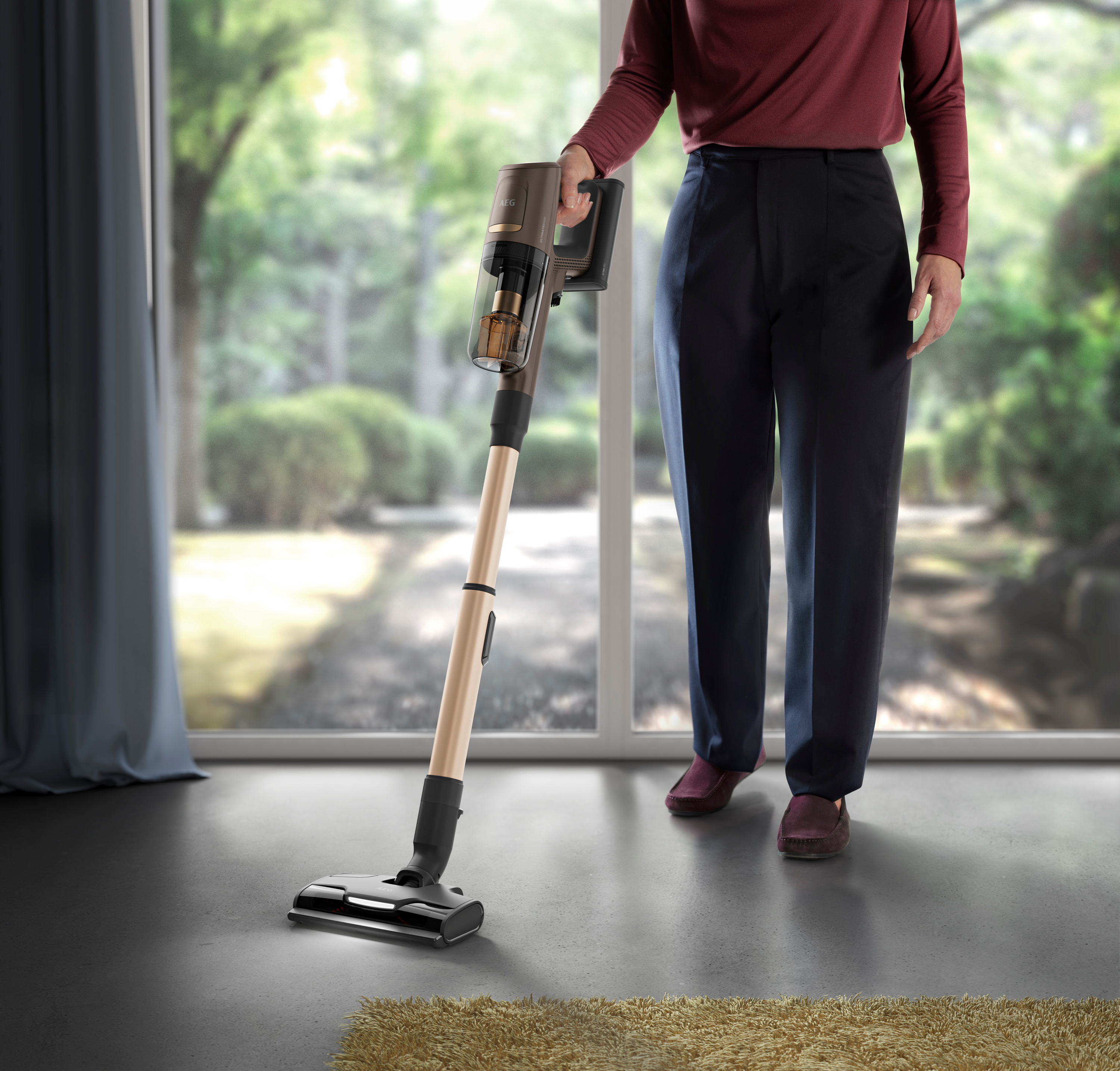 Person verwendet einen gold-schwarzen Staubsauger auf grauem Boden. Im Hintergrund Fenster und Teppich.