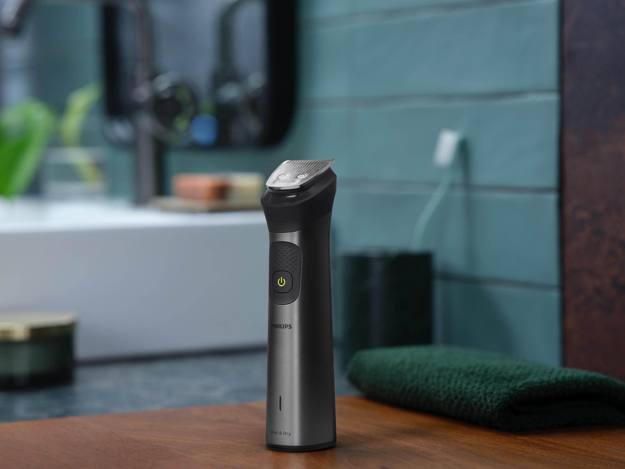 Ein schwarz-silberner Philips Barttrimmer auf einer Holzoberfläche, neben einem grünen Handtuch und einem Glas.