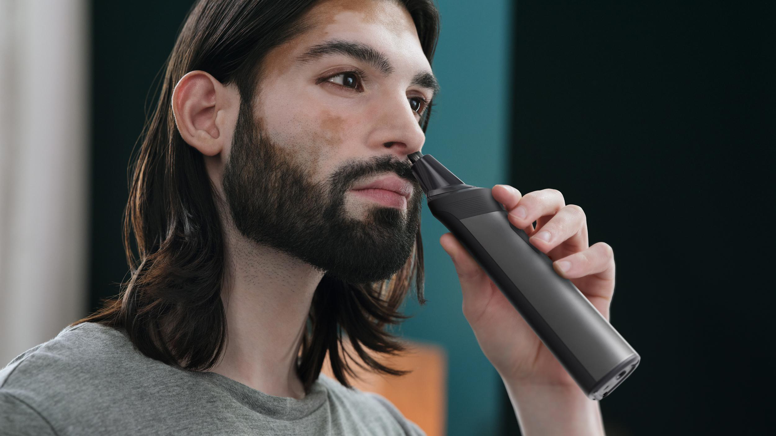 Ein Mann trimmt seine Nasenhaare mit einem schwarzen Trimmer. Hintergrund: türkisfarbene Wand.