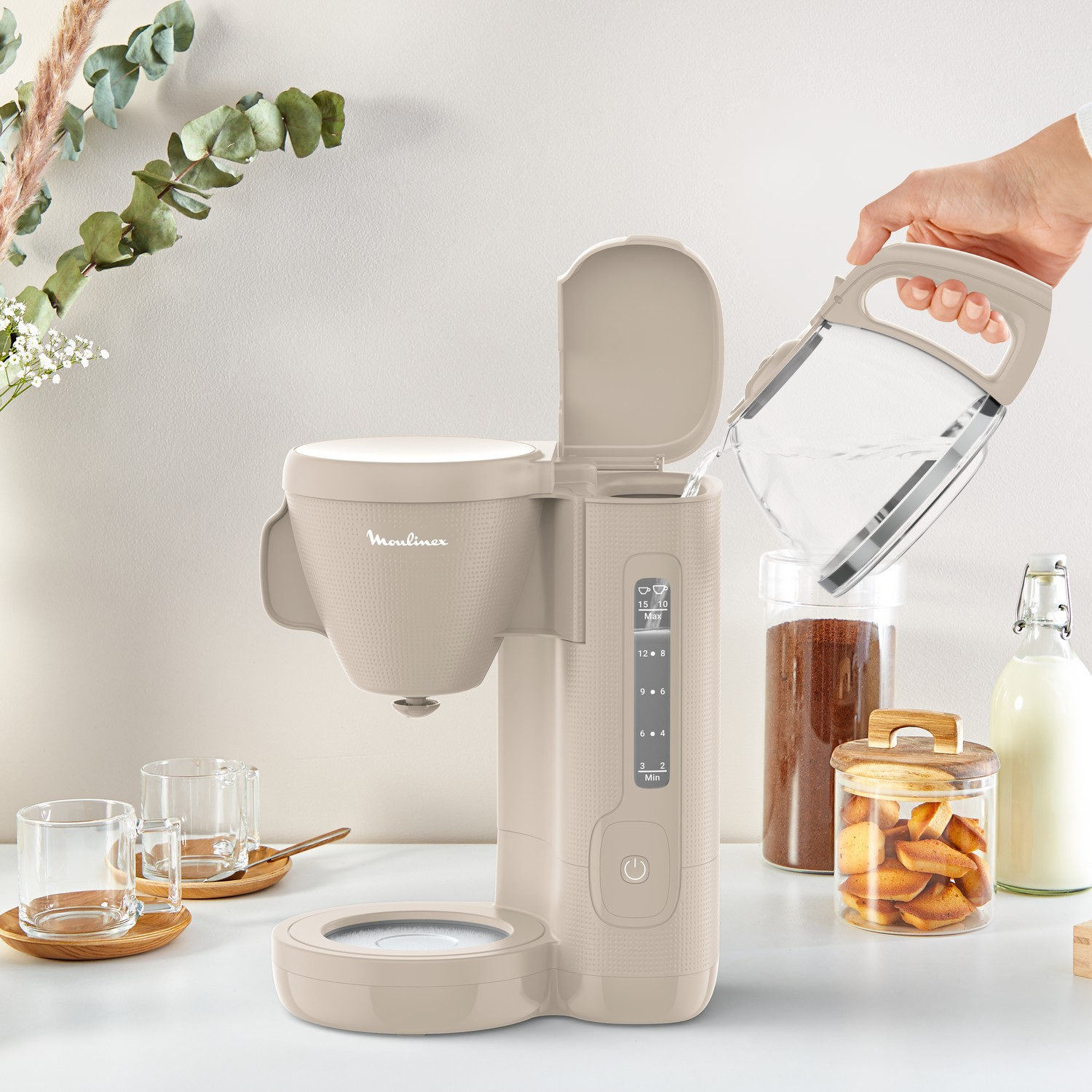 A beige Moulinex coffee machine with a water pitcher, coffee beans, and glasses on a white surface.