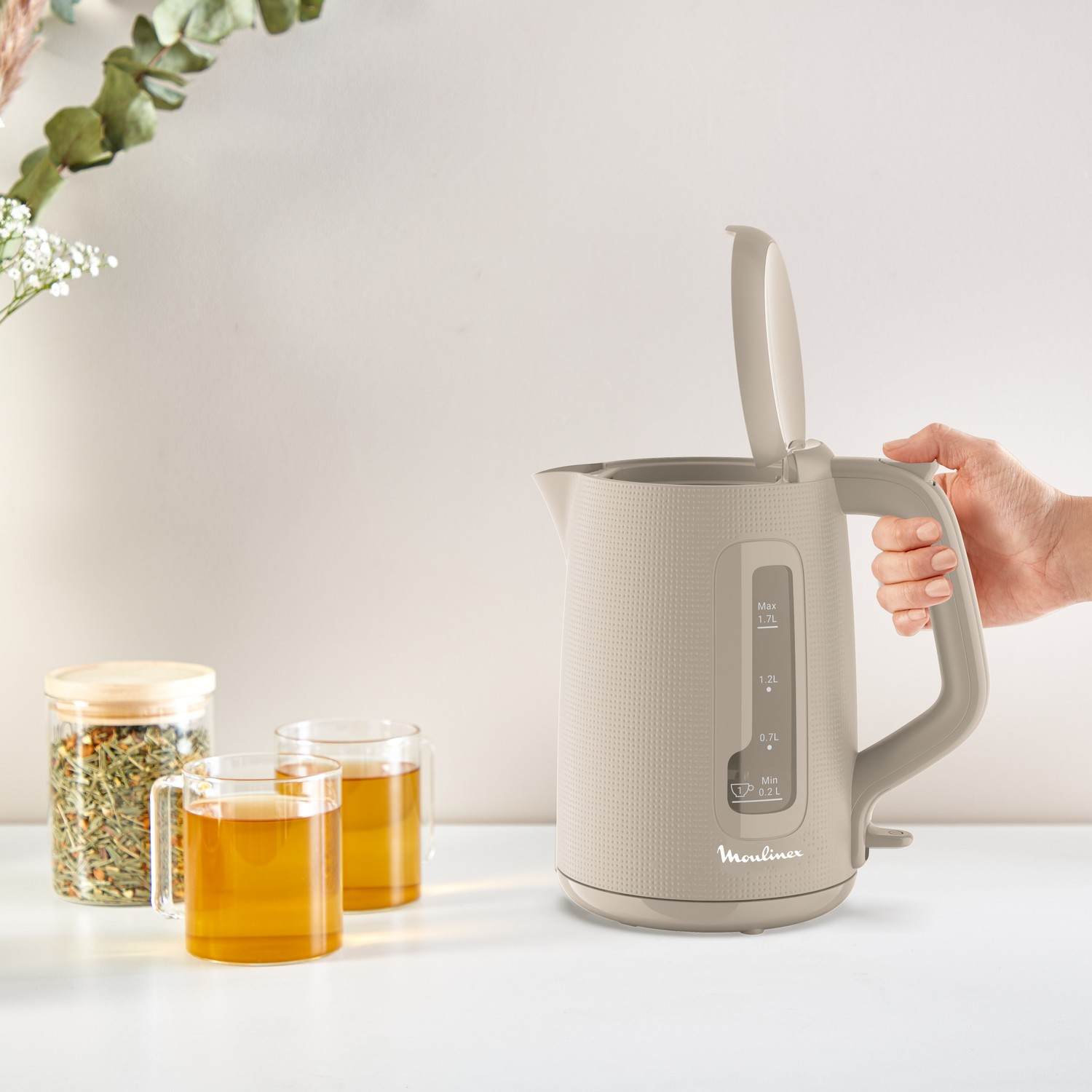 Beige electric kettle with an open lid, next to two glasses of tea and a jar with tea leaves.