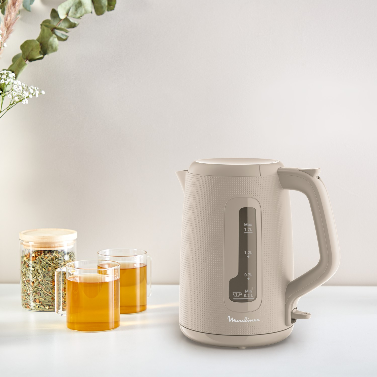A beige electric kettle with tea and glasses on a table, and flowers in the background.
