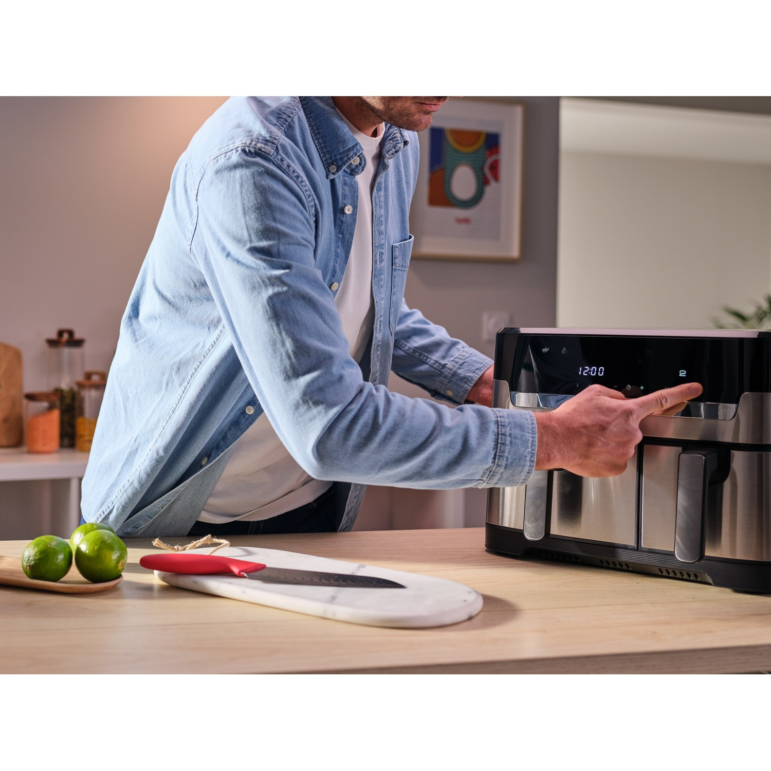Une personne utilise une friteuse à air sur une table en bois, avec des citrons verts, un couteau et une planche à découper.