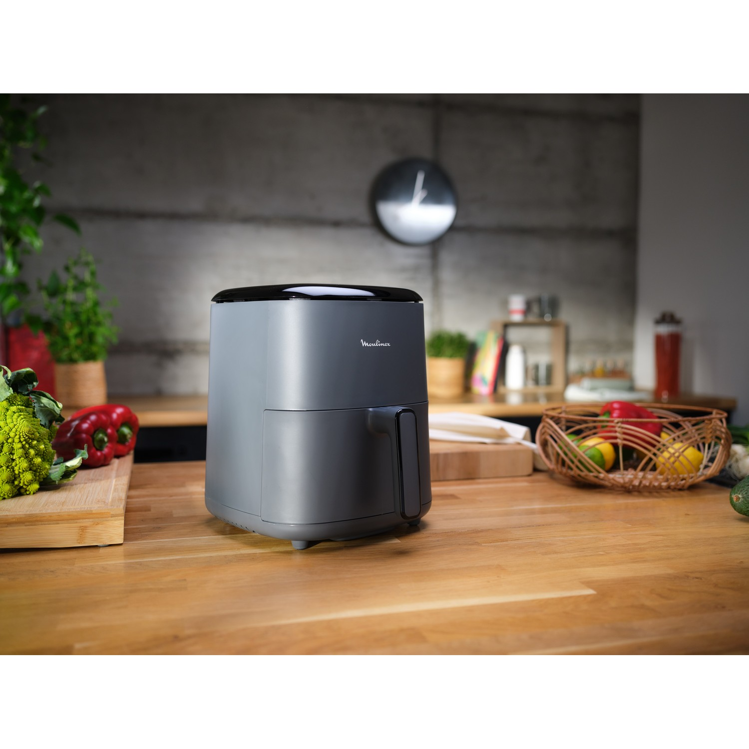 Een grijze airfryer staat op een houten aanrecht met groenten en een klok op de achtergrond.