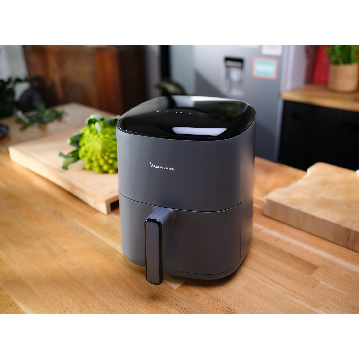 Een grijze airfryer op een houten aanrecht. Een groen object en een snijplank staan op de achtergrond.