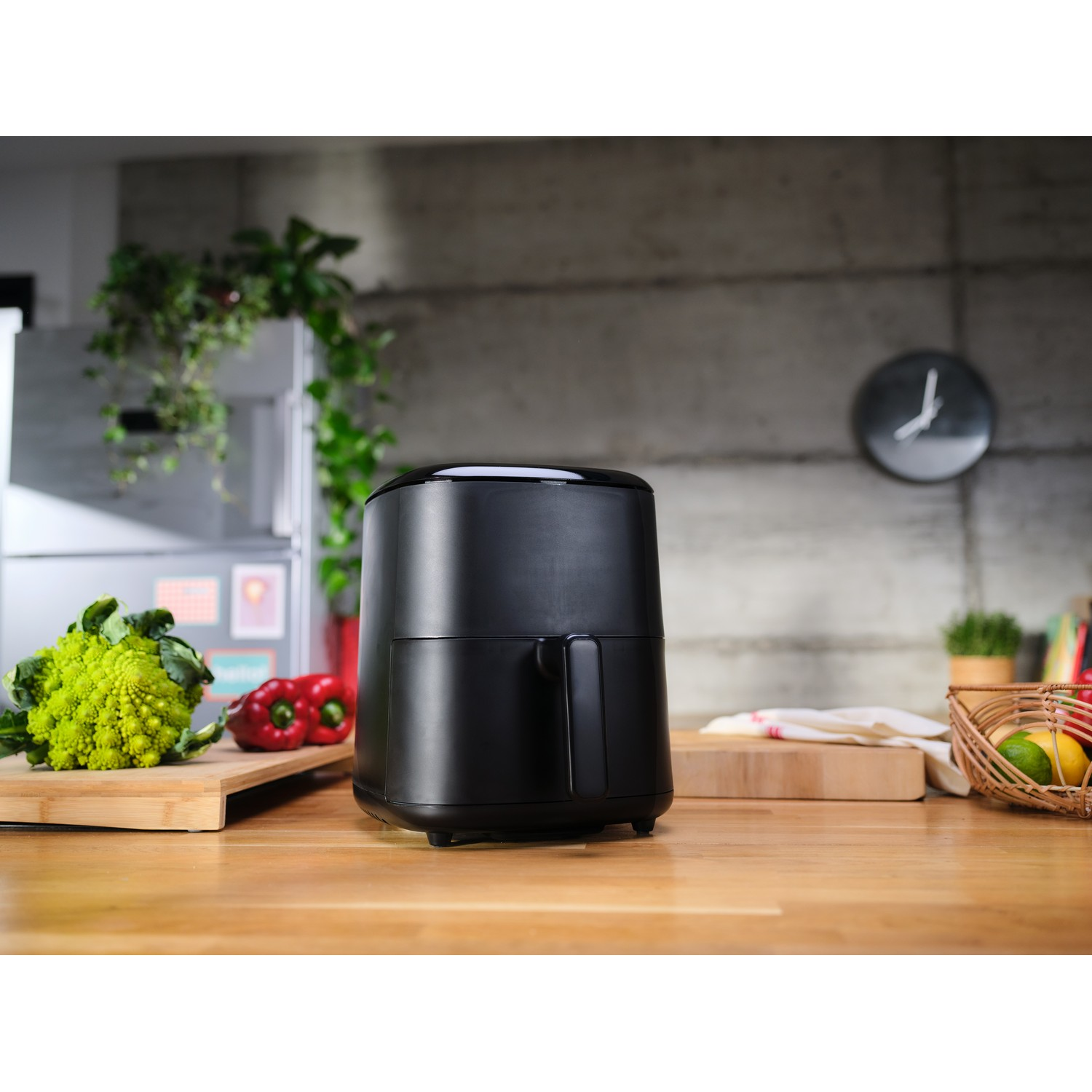 Zwarte airfryer op een houten aanrecht, groenten en klok op de achtergrond.