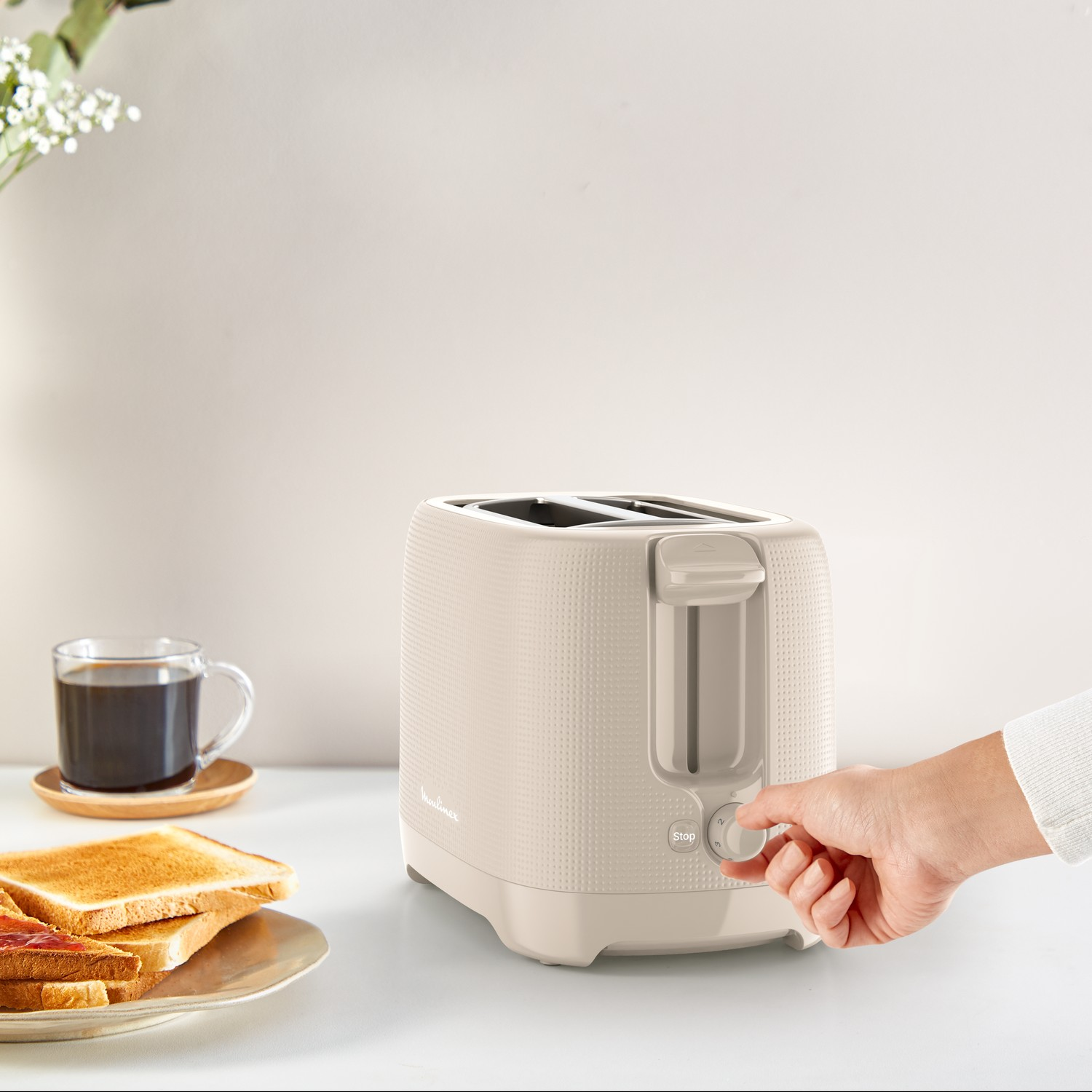 A toaster. A hand is turning a knob. Toast and coffee on the table.