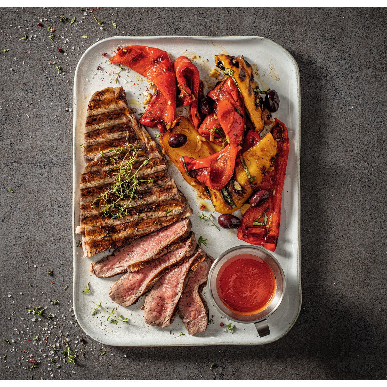 A plate with grilled steak, roasted peppers, olives, and a small bowl of sauce. Garnished with herbs.