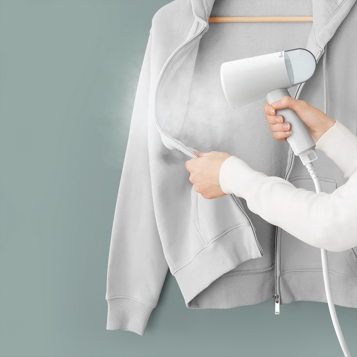 Een persoon stoomt een grijze hoodie met een witte handheld steamer. De muur is blauwgroen.