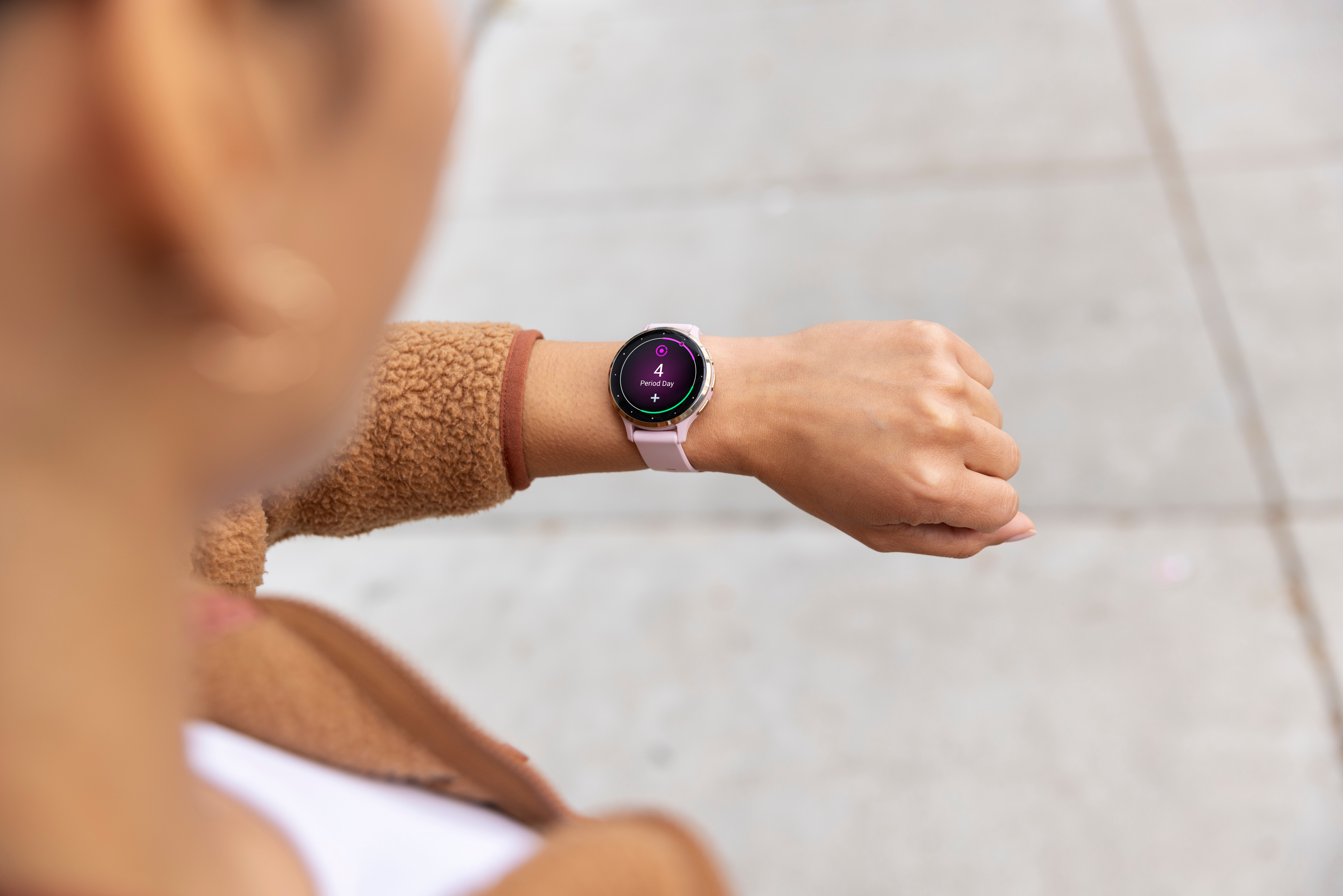 Das Handgelenk einer Person mit Smartwatch zeigt den Tag 4 der Periode an. Die Person trägt eine braune Jacke.
