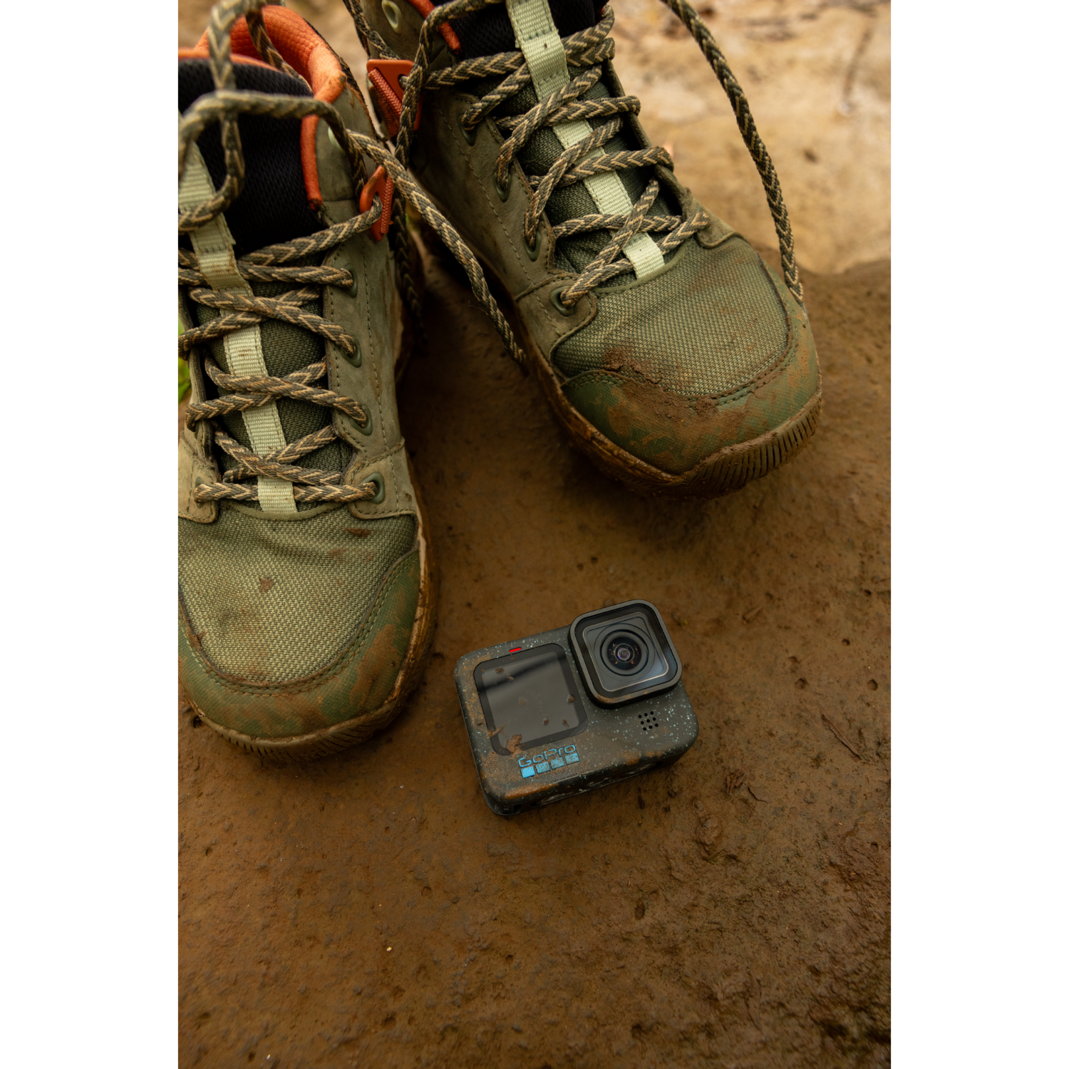 Pair of green hiking boots and a black GoPro camera on a wet brown surface. Boots are muddy.