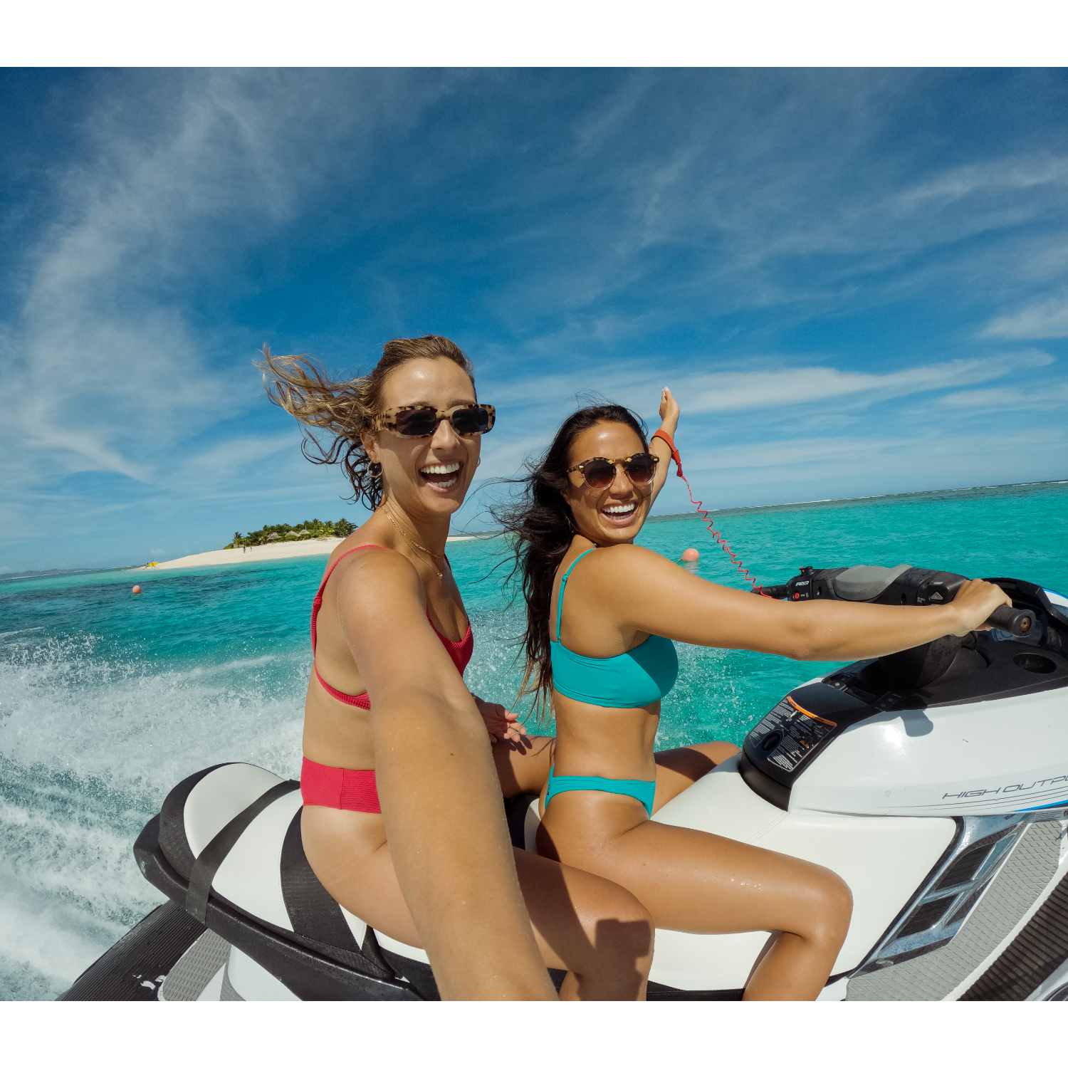 Two women on a jet ski, one waving, with blue water and sky.