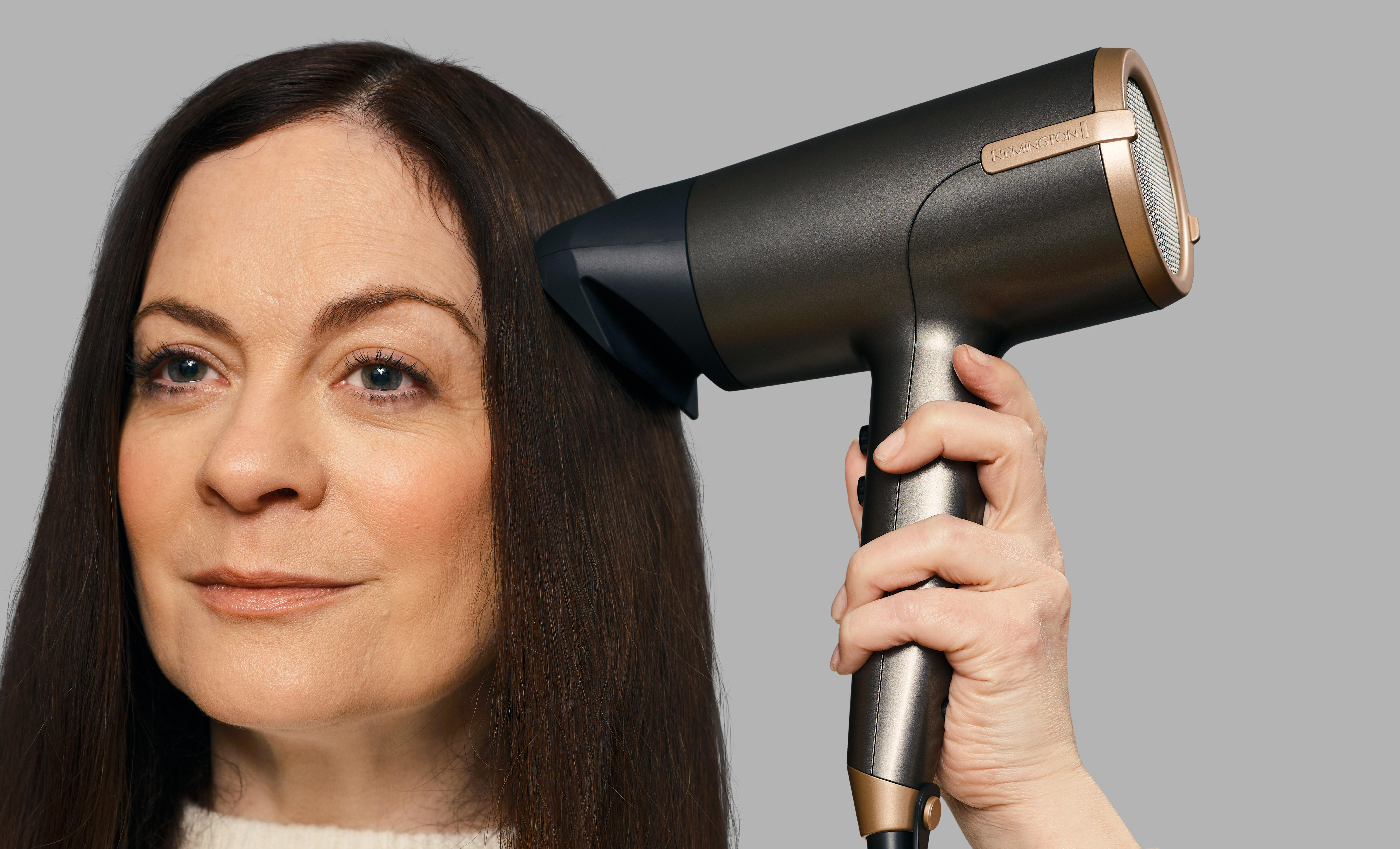 Frau, die einen Haartrockner für ihr Haar benutzt. Grauer Hintergrund. Sie trägt einen Pullover.