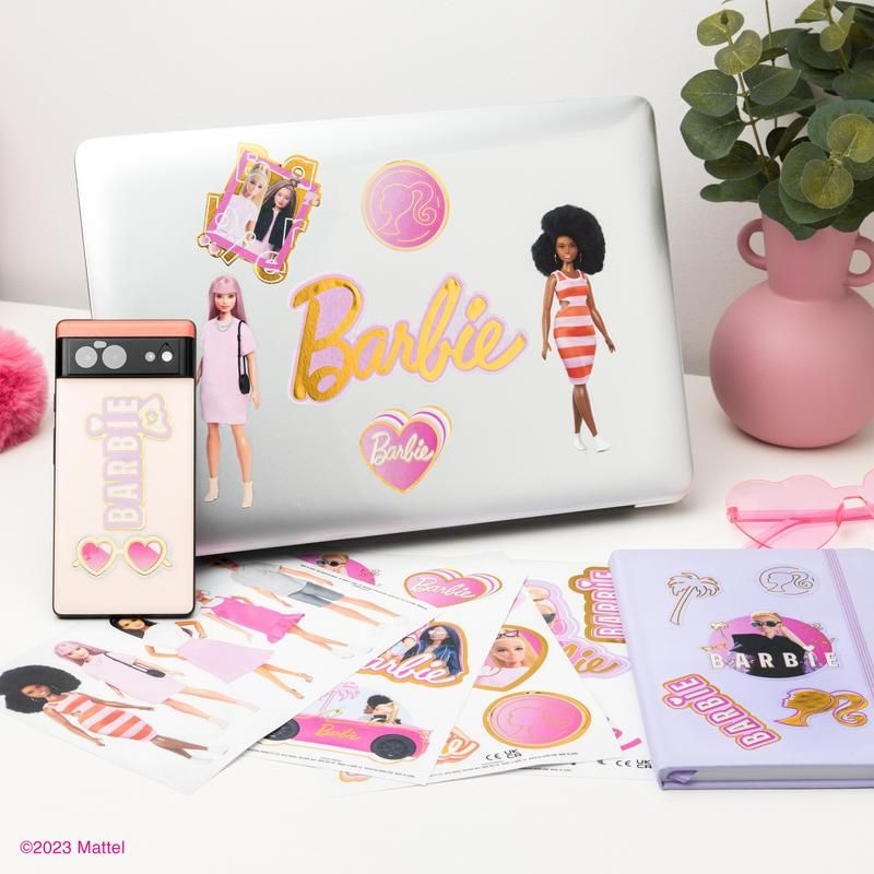 Egy Barbie matricákkal ellátott laptop, telefon, babák, egy rózsaszín váza zölddel és egy jegyzetfüzet fehér felületen.