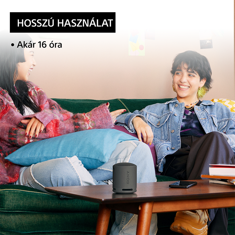 Két nő ül egy zöld kanapén, egyikük egy kis szürke hangszórót használ. Szöveg: hosszú használat.