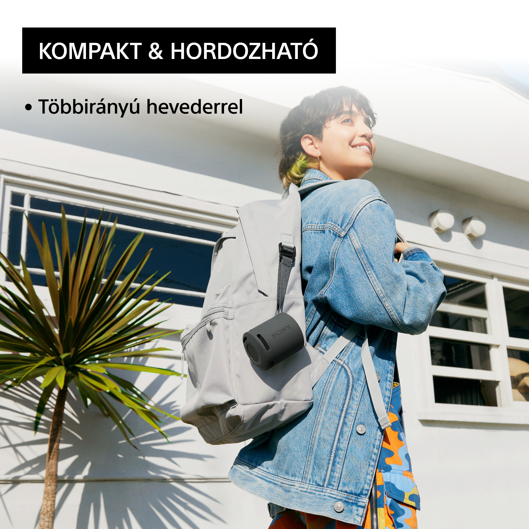 Egy nő hátizsákkal, fekete hangszórót visz, egy pálmafával rendelkező ház előtt állva.
