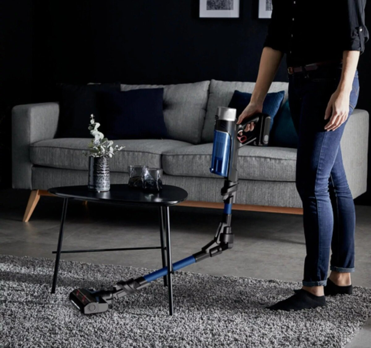 Osoba odkurzająca pokój odkurzaczem bezprzewodowym. Szara sofa i stół w tle.