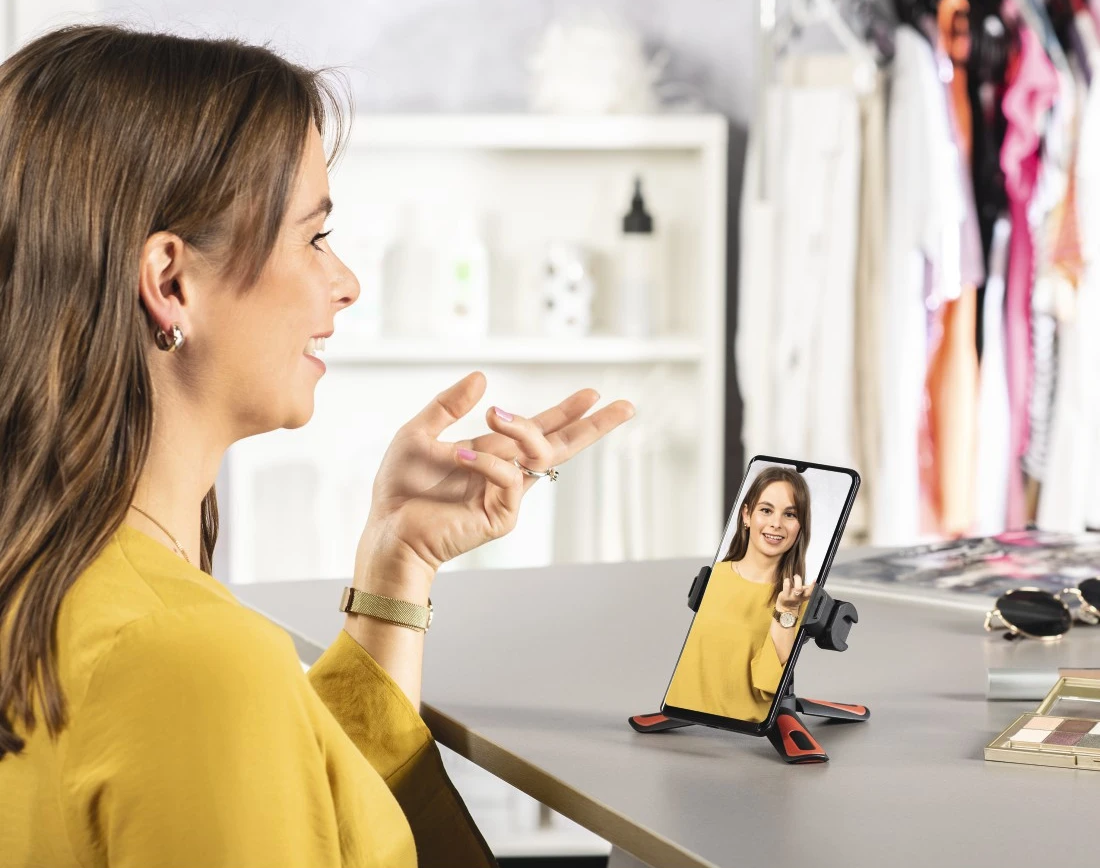 Sárga felsőt viselő nő videohívást folytat, telefonnal egy állványon.