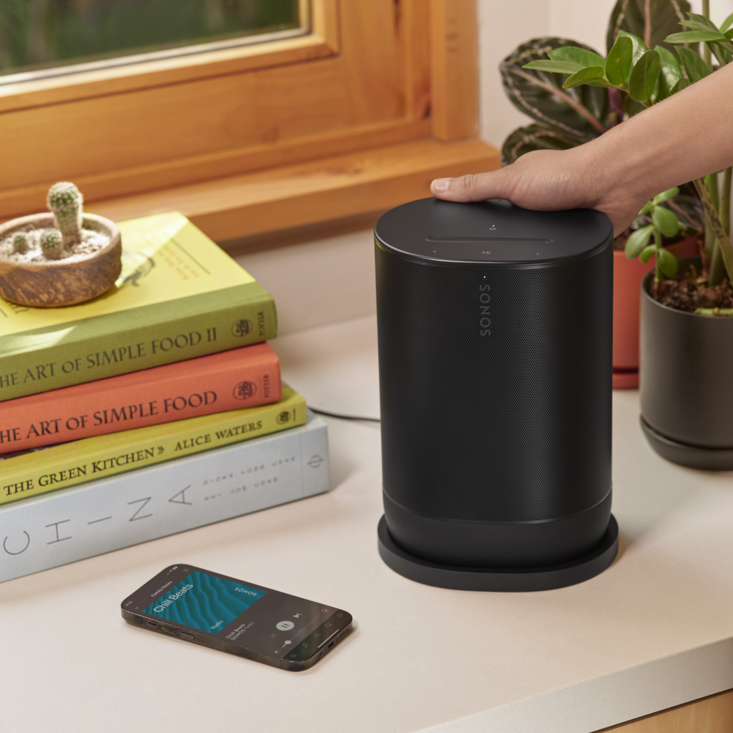 Zwarte Sonos-luidspreker met een hand erop, boeken en een telefoon ernaast.