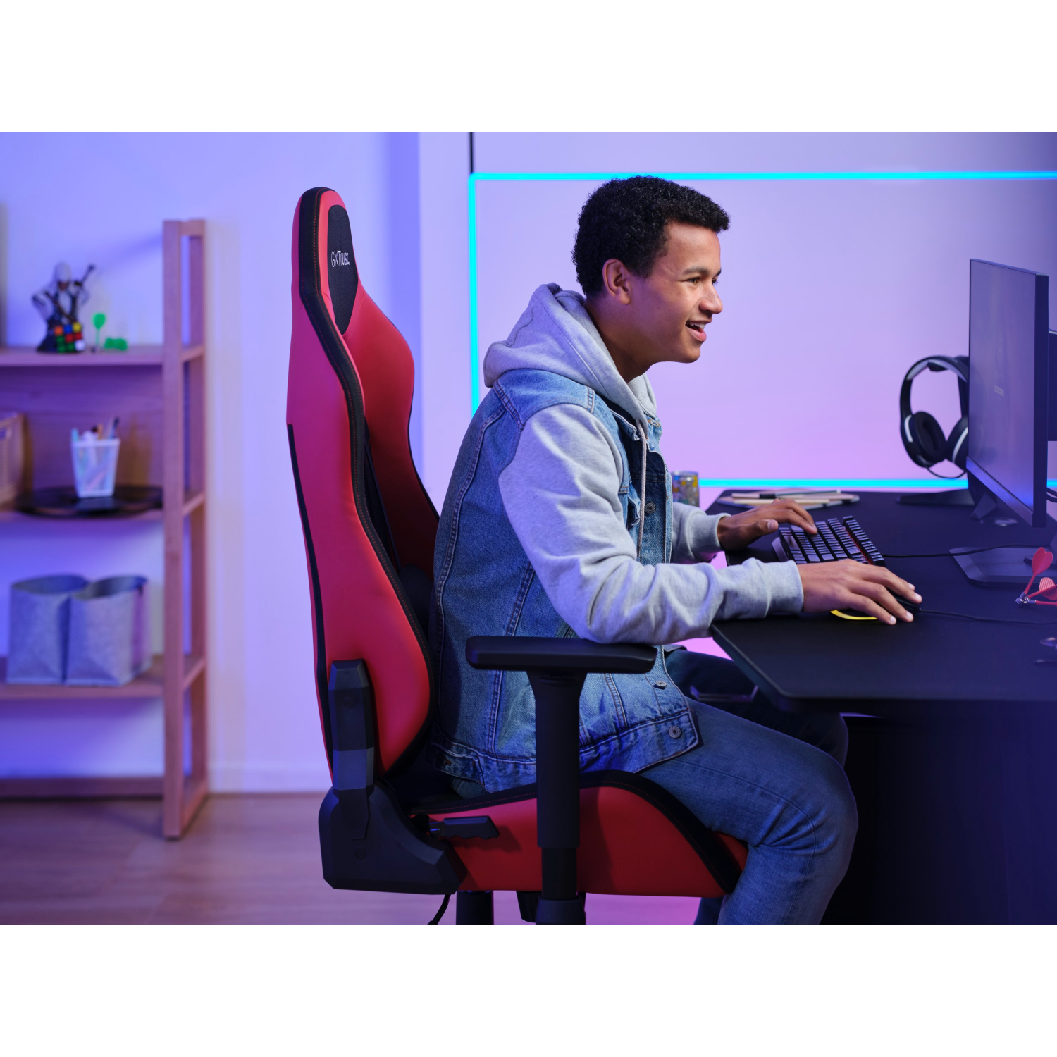 Een jonge man is aan het gamen, zit in een rode stoel, kijkt naar een computerscherm.