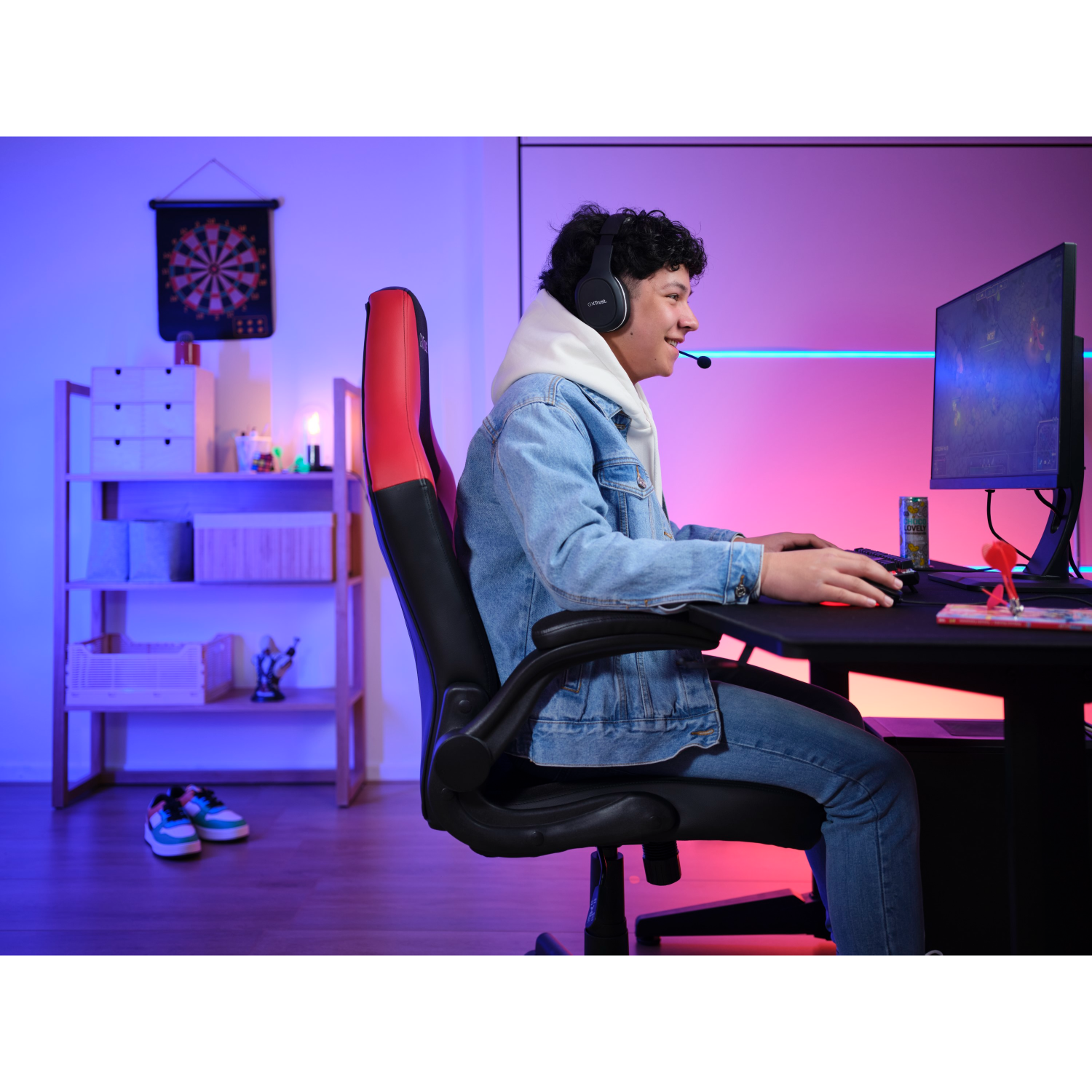 A gamer wearing headphones plays a game on a computer with a red gaming chair.