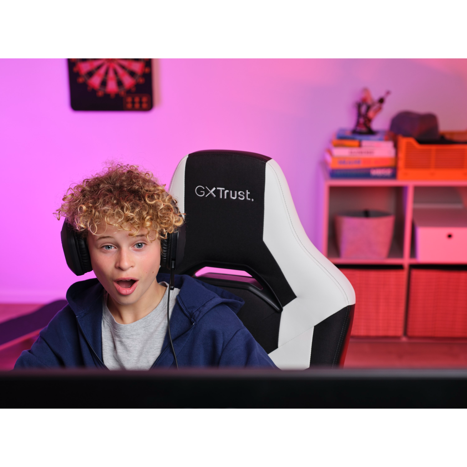 Boy with headphones is surprised. He sits in a gaming chair. Background has pink and white shelves.
