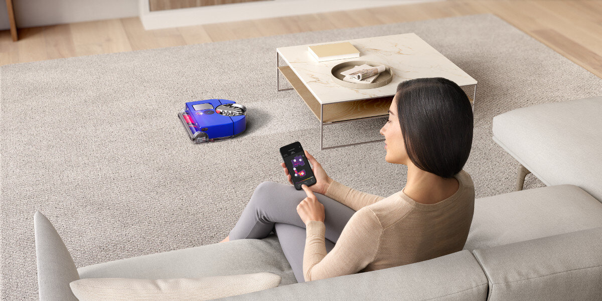 Kobieta na sofie korzystająca ze smartfona. Robot odkurzający czyści dywan.