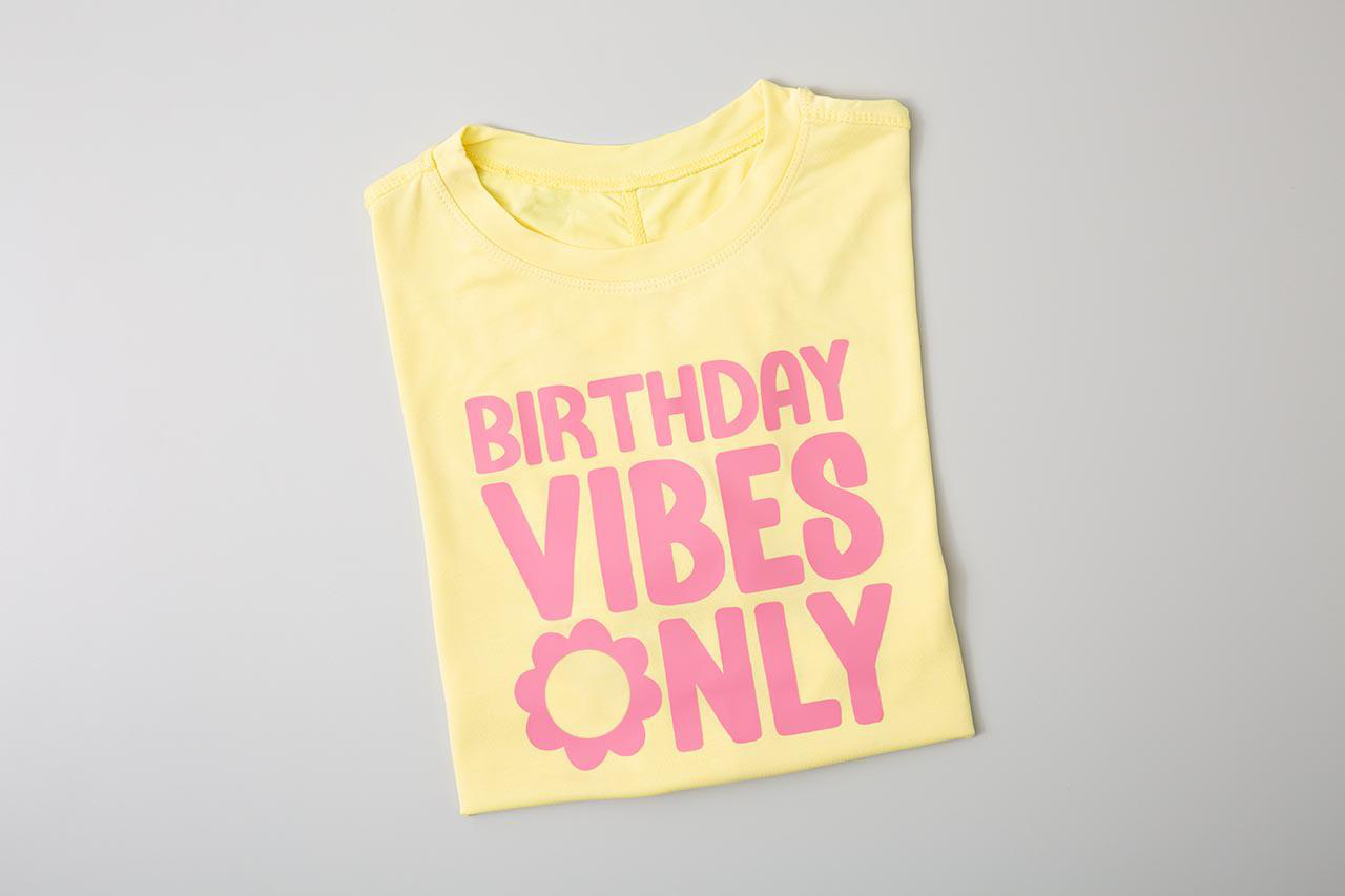 Gelbes T-Shirt mit pinkfarbenem Text 'BIRTHDAY VIBES ONLY' und einer Blume. Liegt flach auf grauer Oberfläche.
