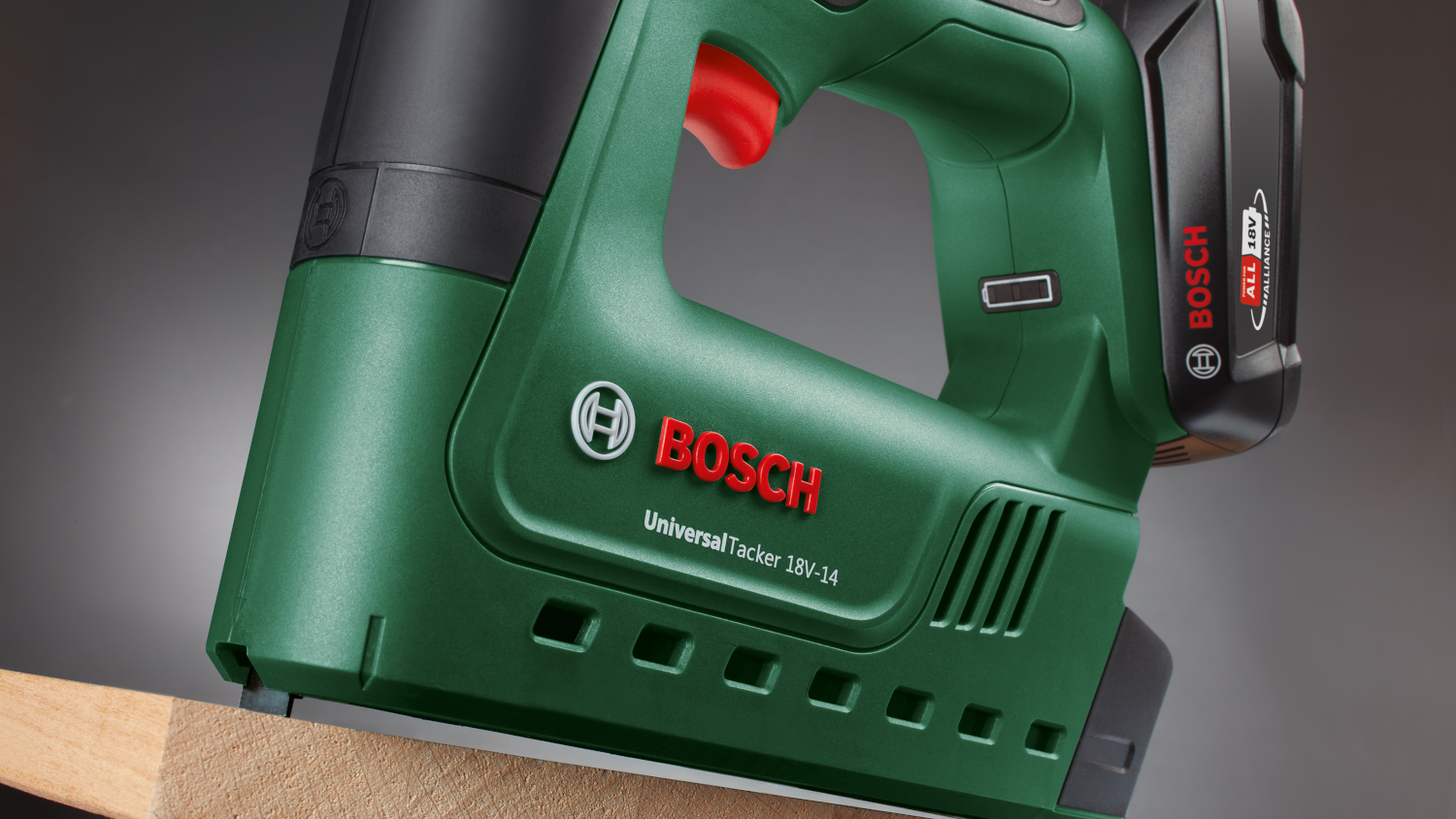 Zöld Bosch Universal Tacker 18V-14 tűzőgép fán. Piros ravasz. Fekete fogantyú és akkumulátor. Szürke háttér.