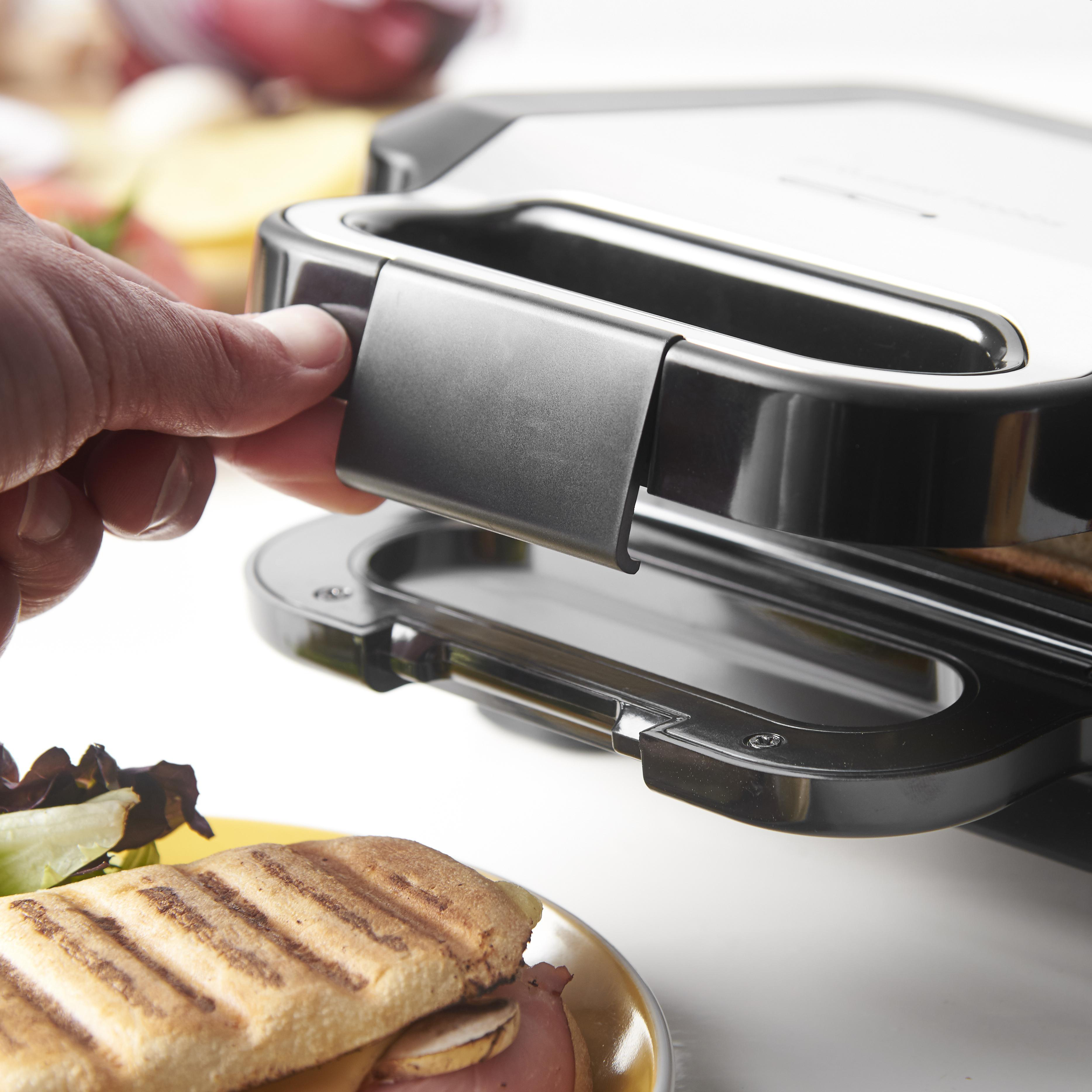 Eine Person schließt einen Sandwichgrill. Ein Sandwich mit Schinken und Käse liegt auf einem gelben Teller.