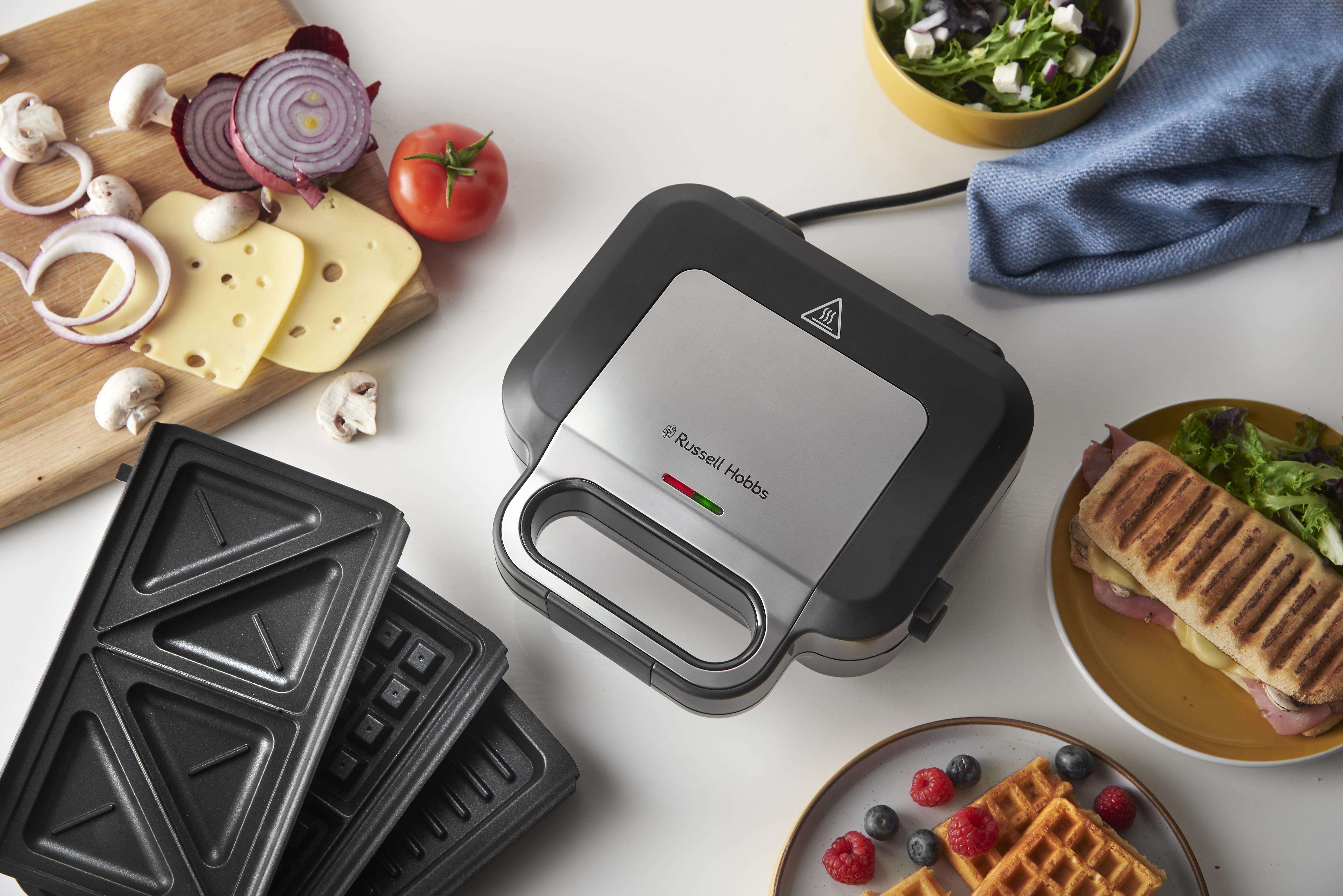 Sandwichmaker mit Waffeln, Zutaten und Schneidebrett auf einem Tisch.