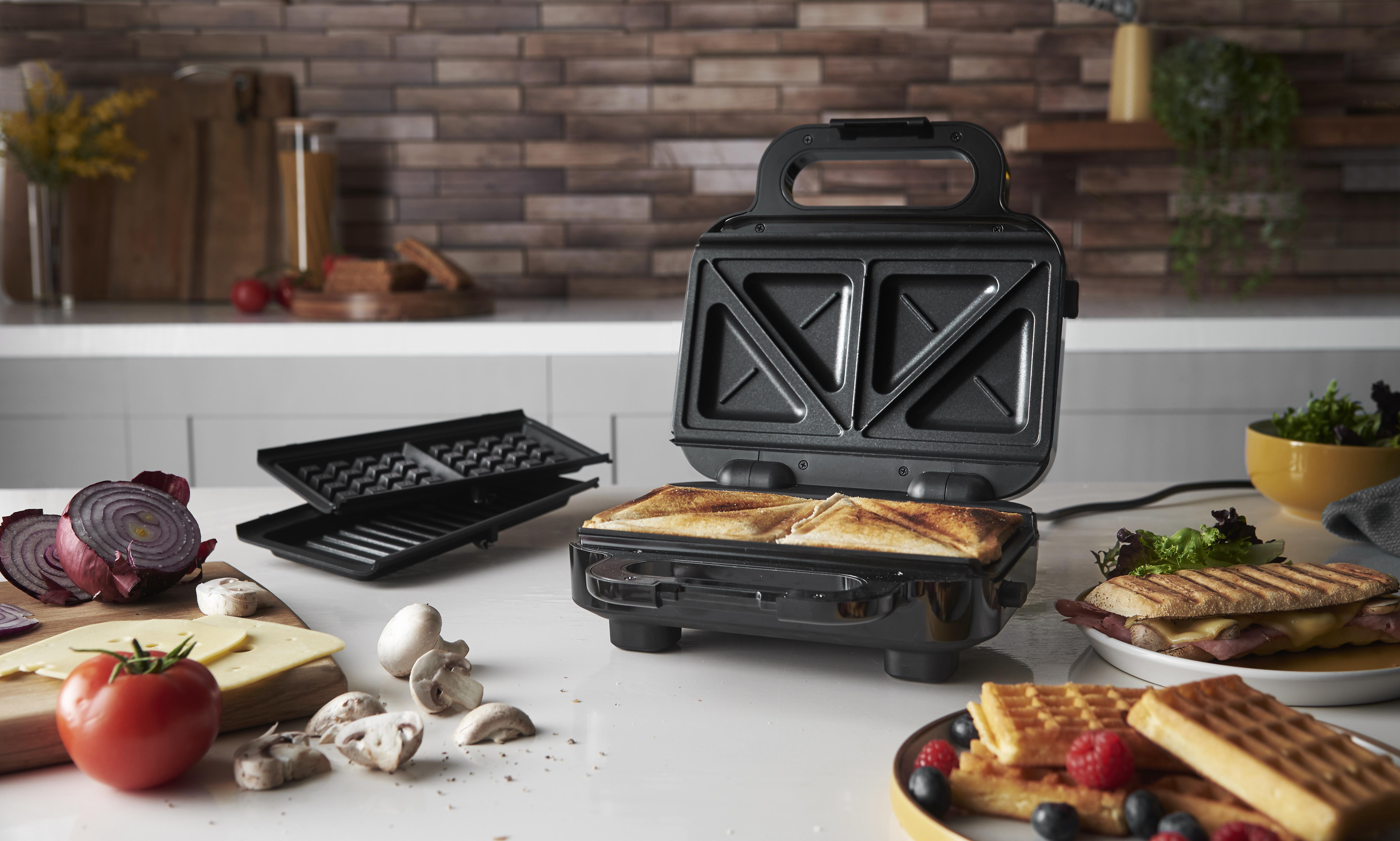 Sandwichmaker mit Sandwiches und Waffeln auf weißer Oberfläche. Gemüse und Zutaten daneben.