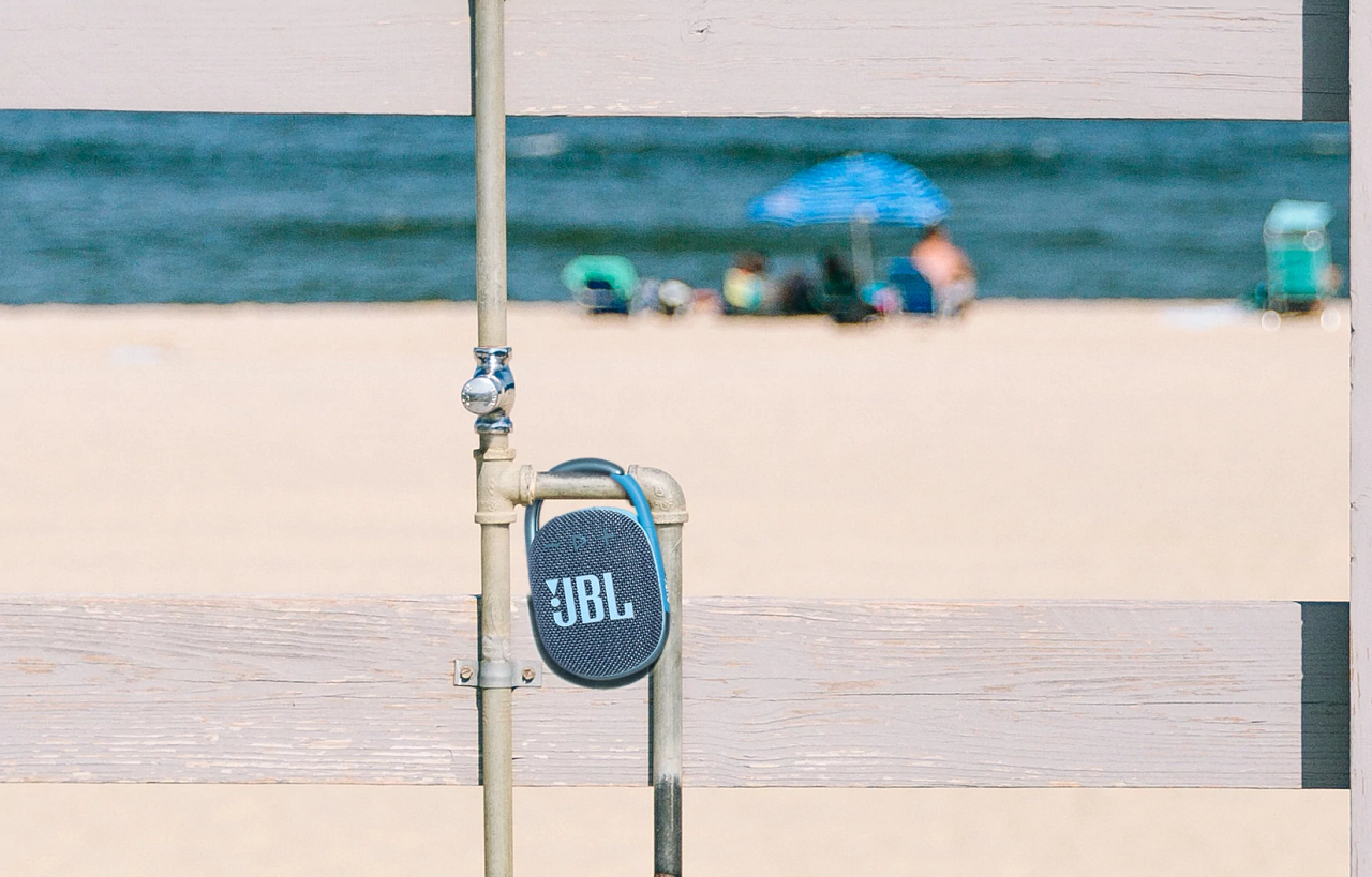 Ein blauer JBL-Lautsprecher hängt an einem Metallrohr. Im Hintergrund eine Strandszene.
