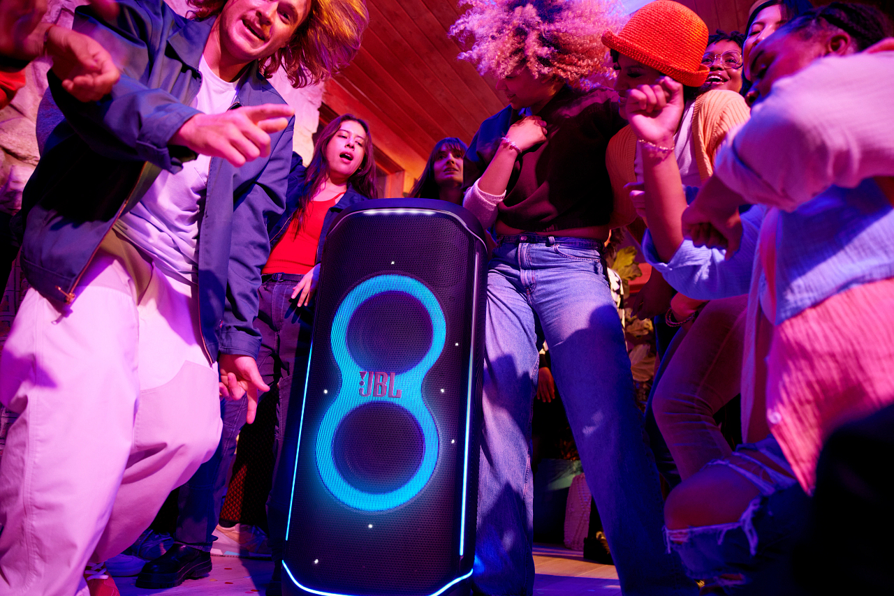 Leute tanzen um einen JBL-Lautsprecher. Der Lautsprecher hat leuchtend blaue Akzente. Die Szene ist in Rosa- und Lilatönen gehalten.
